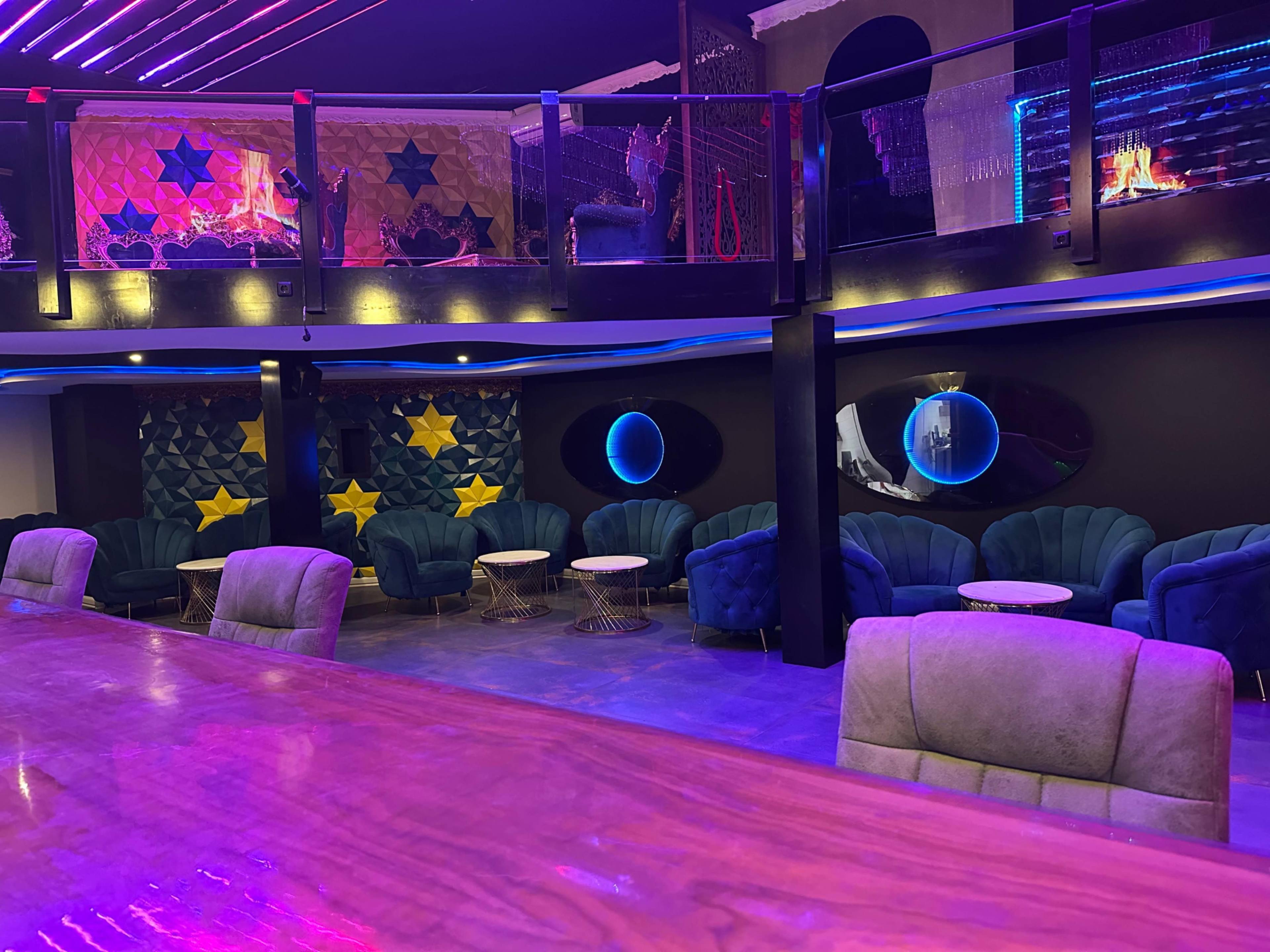 The image shows a dimly lit lounge with modern seating arrangements, colorful lighting, and a bar area in the foreground.