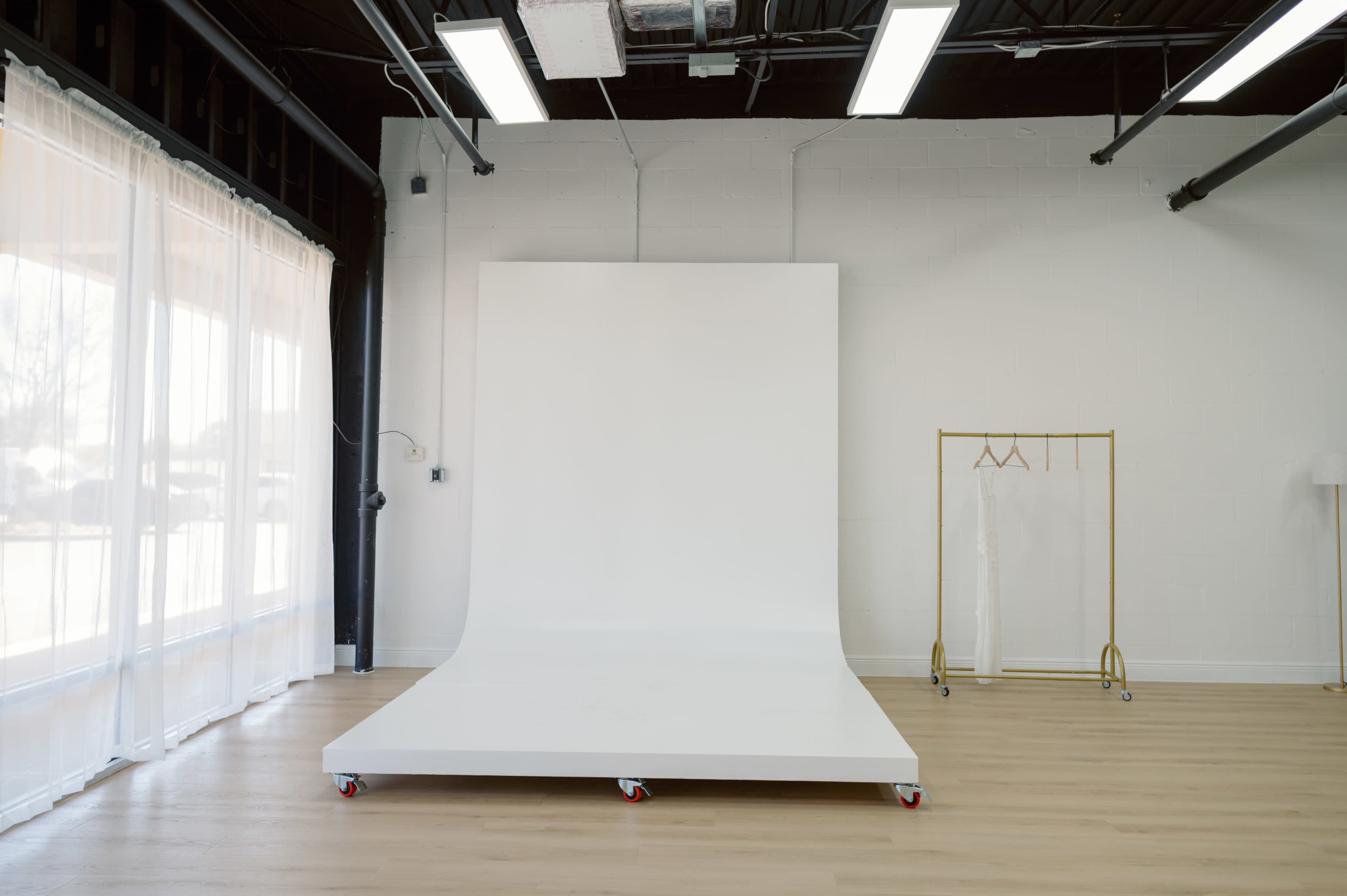 The image shows an empty photography studio with a white backdrop and a clothing rack on wheels against a bright, spacious interior.