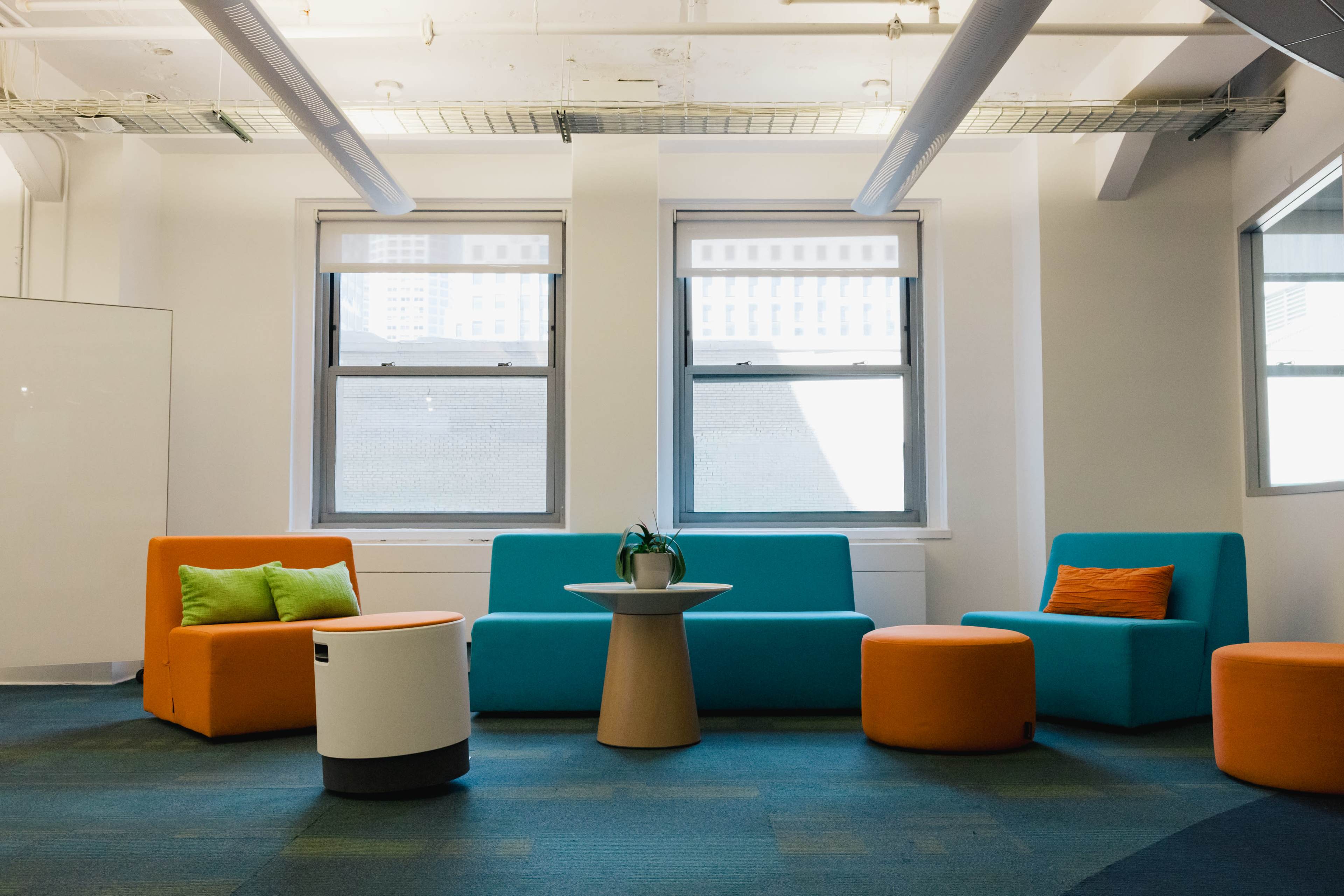 The image shows a brightly colored modern lounge area featuring orange and blue seating arrangements along with a small wooden table and green plant decor.