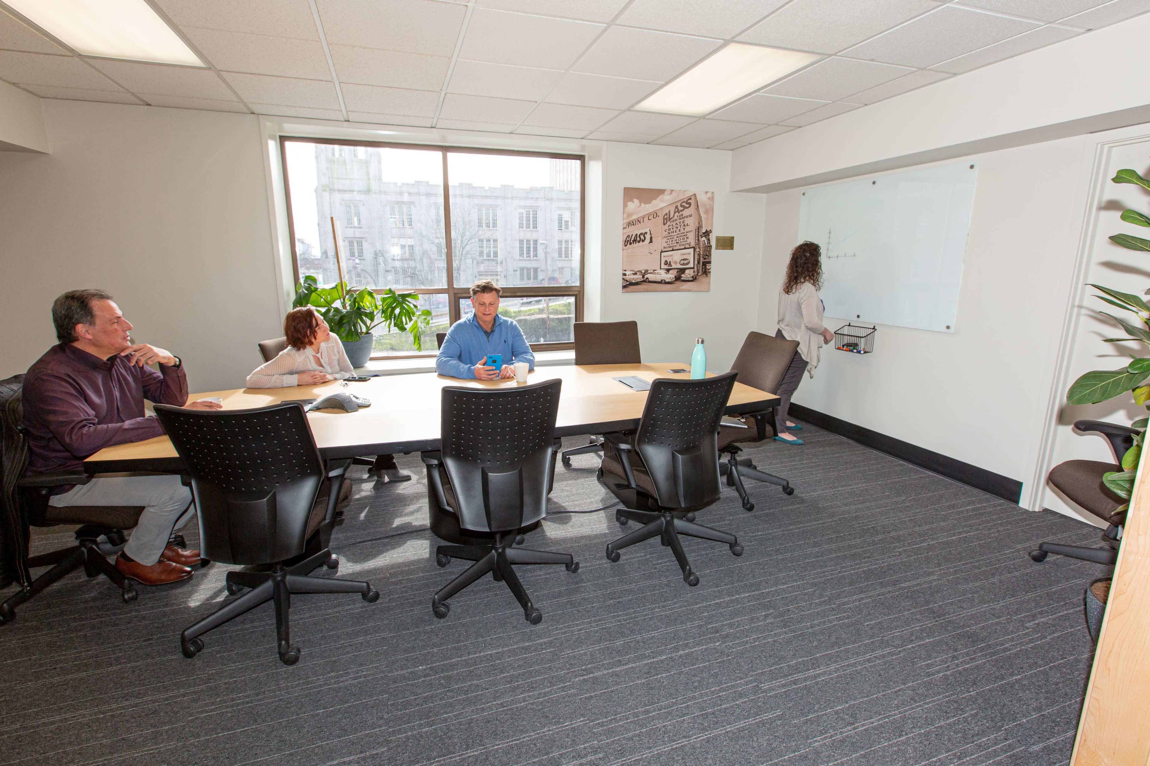 A group of four people is seated at a conference table in a well-lit meeting room, with one person writing on a whiteboard while others observe.