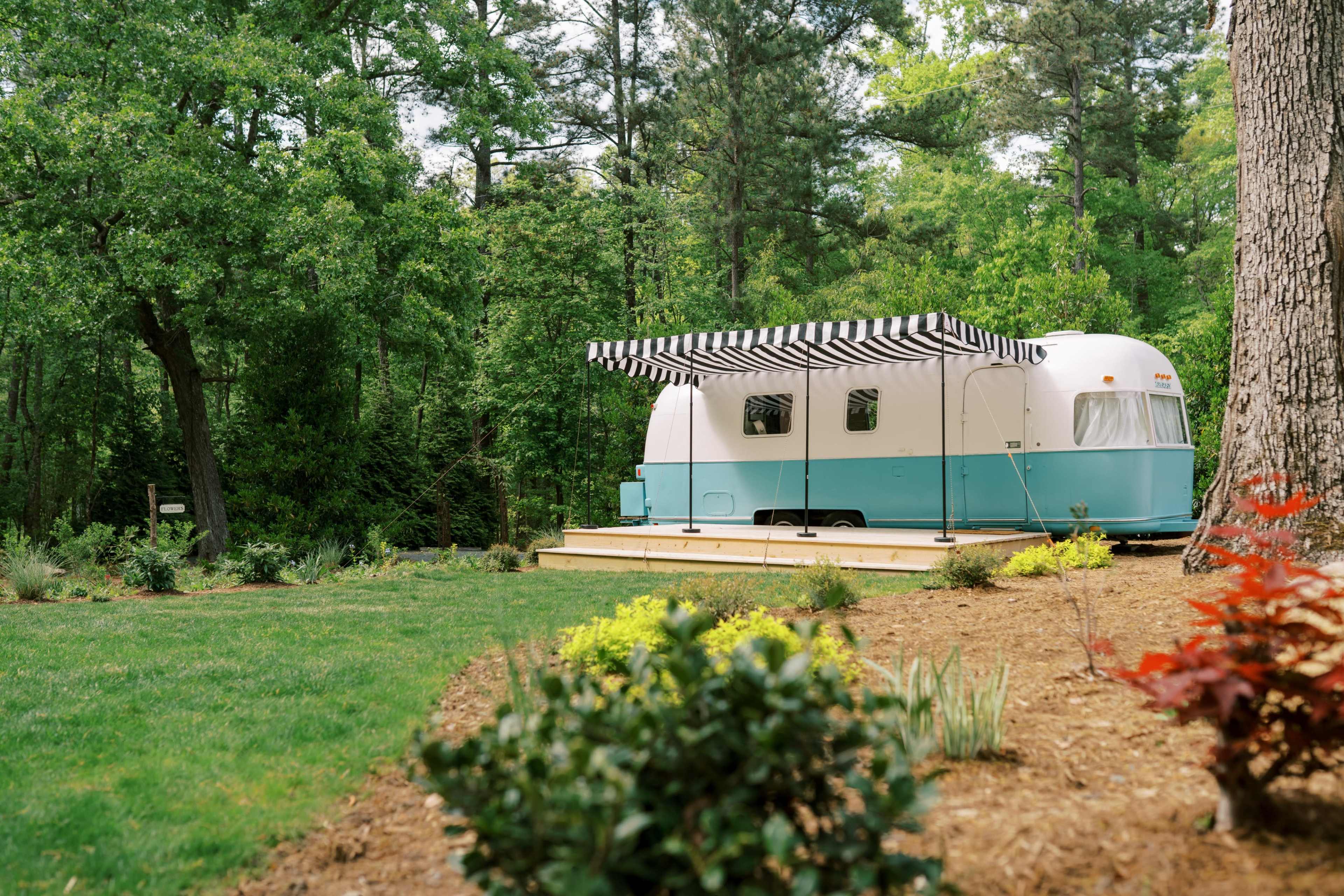 A vintage Airstream trailer with a striped awning is parked on a wooden deck surrounded by lush greenery and landscaped shrubs.