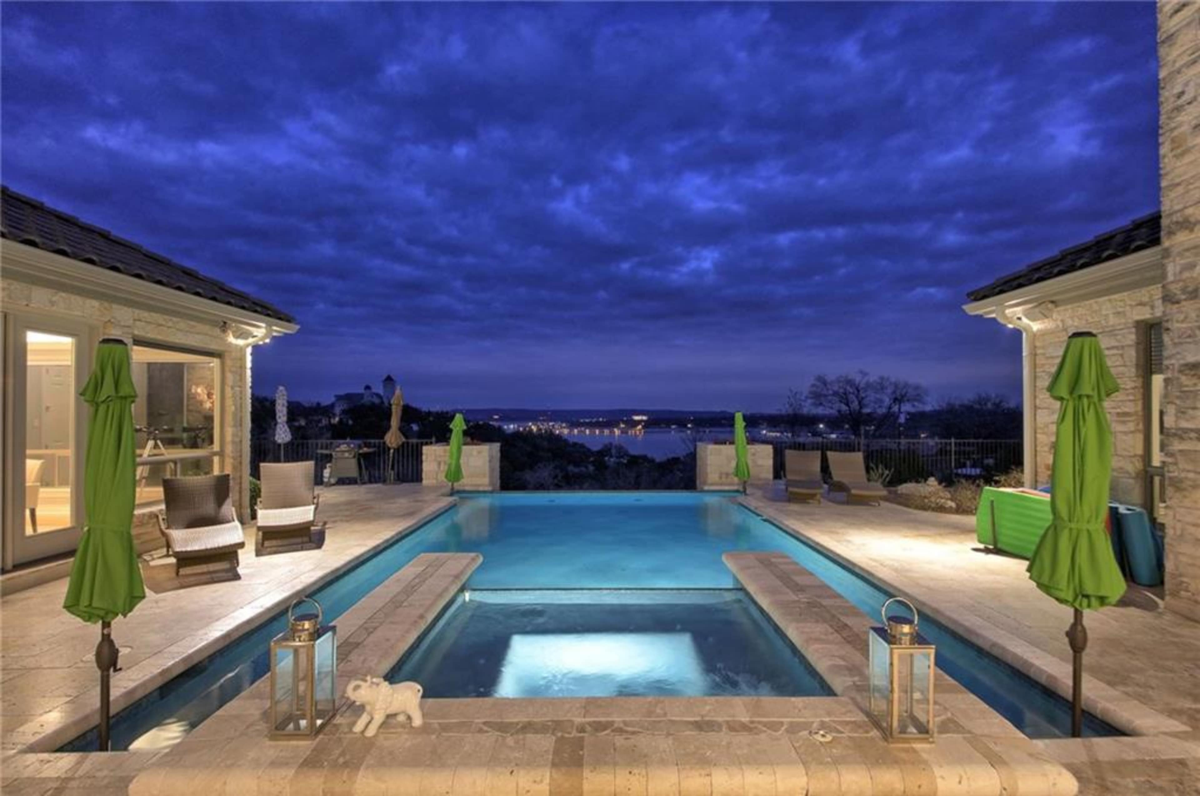 A well-lit pool area features green umbrellas and overlooks a distant skyline under a cloudy twilight sky.