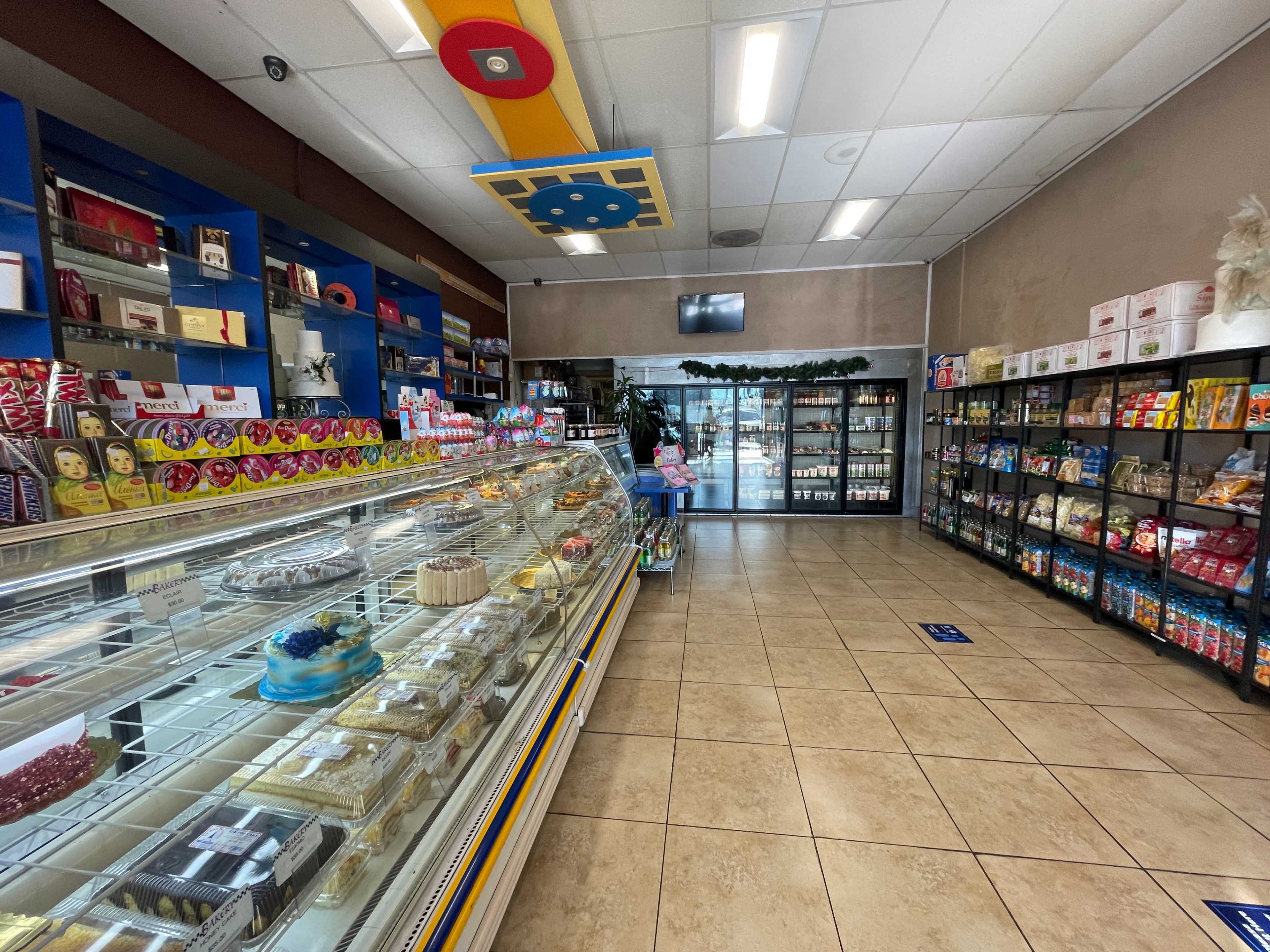 The image shows the interior of a shop featuring a display case with various desserts on the left and shelves stocked with packaged goods on the right.