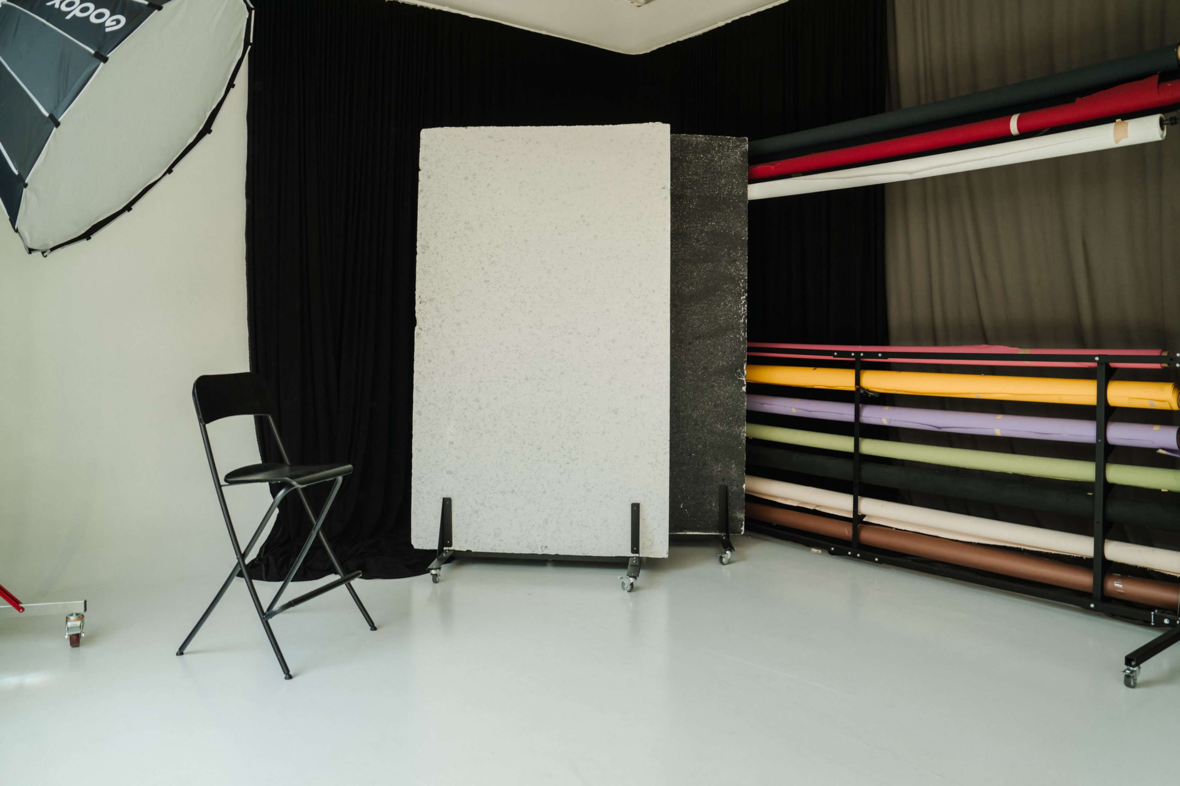 The image shows a photography studio with a black backdrop, a gray foam board set on wheels, a folding chair, and a rack of colored backdrop rolls in the background.