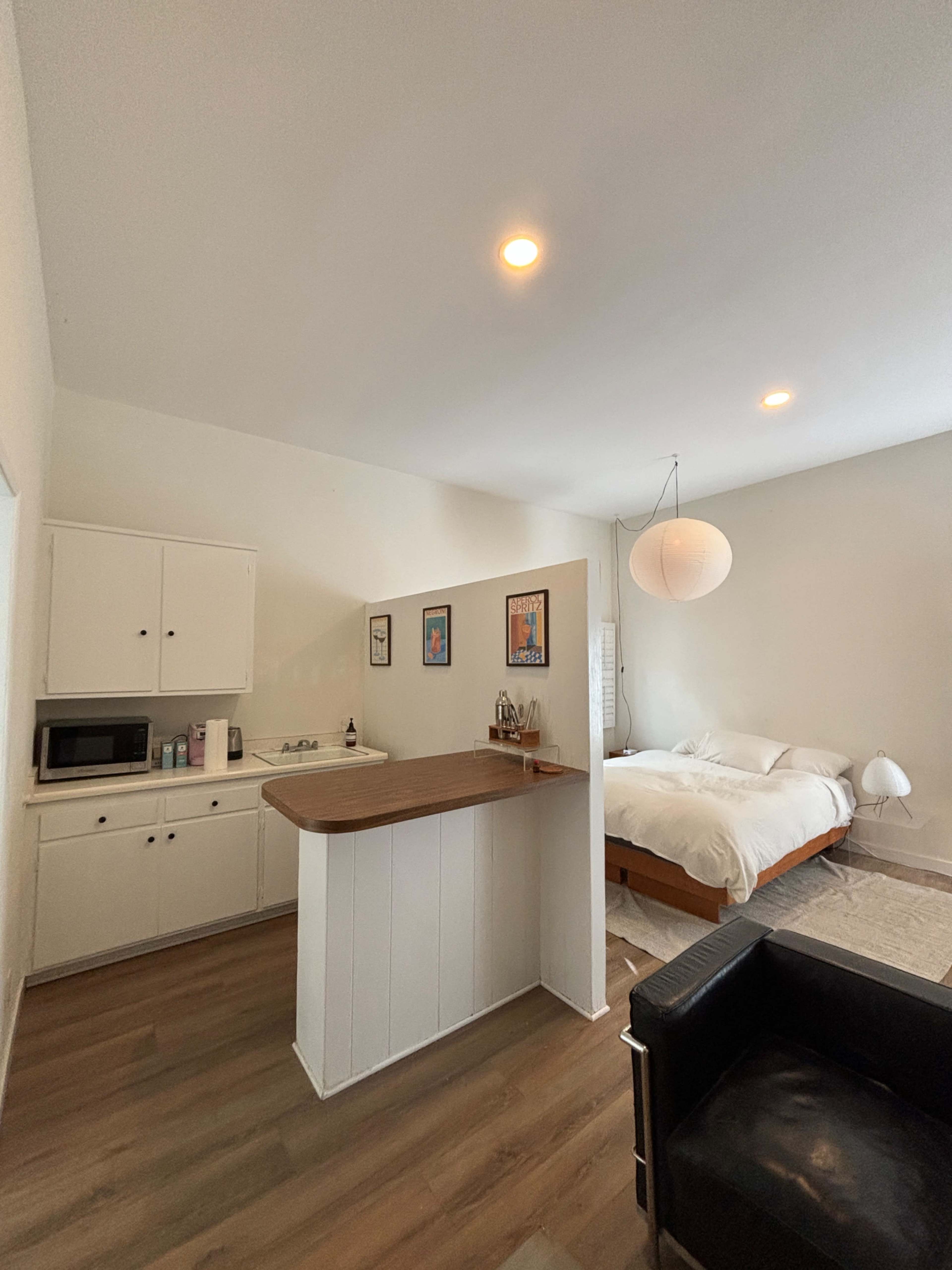 The image shows a small studio apartment with a kitchenette on one side and a bed on the other, featuring a light-colored floor and a modern lamp beside the bed.
