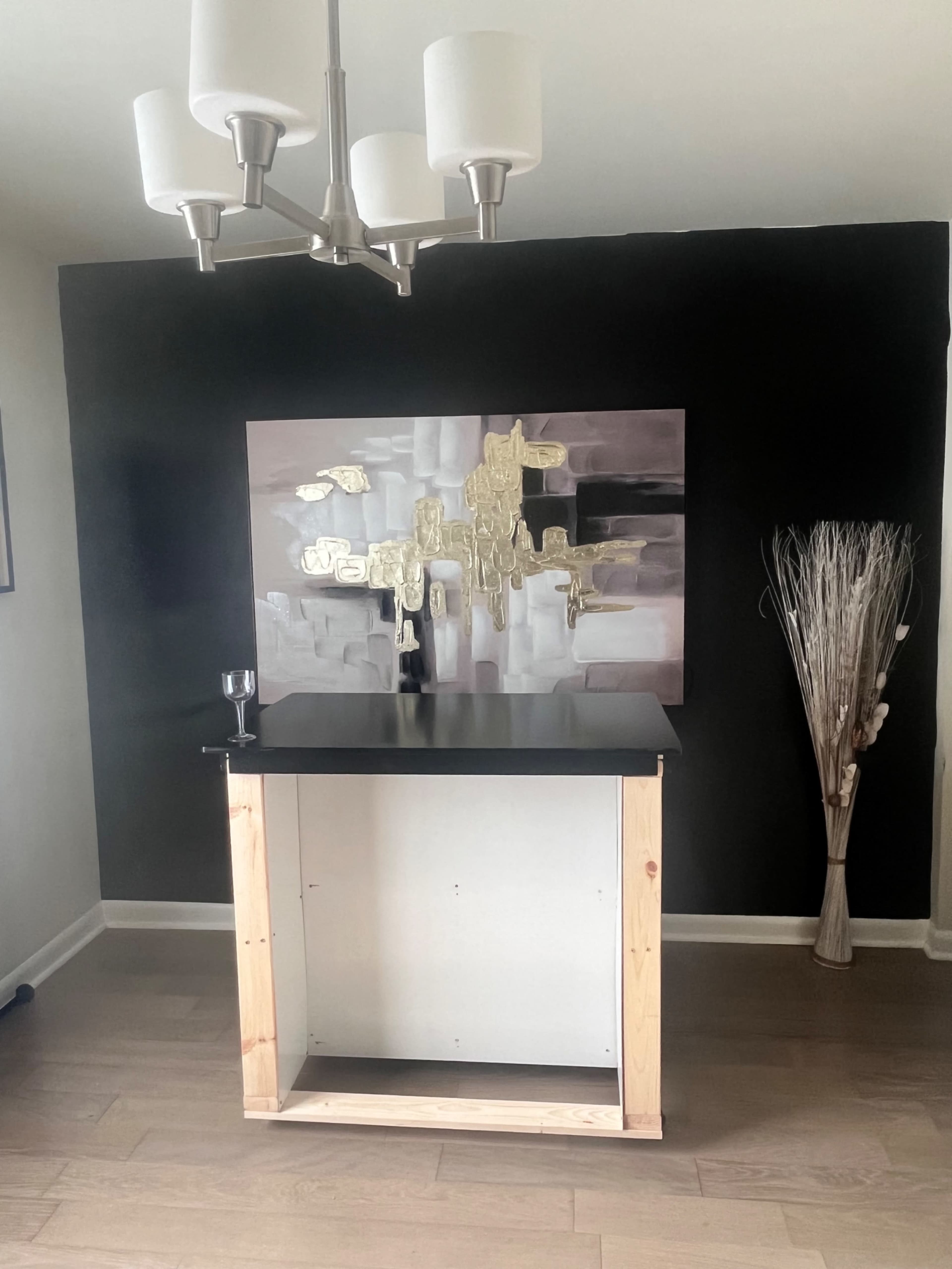 The image shows a minimalist bar area with a black wall, a large abstract painting, a wooden-framed bar counter, and decorative grass in a tall vase.