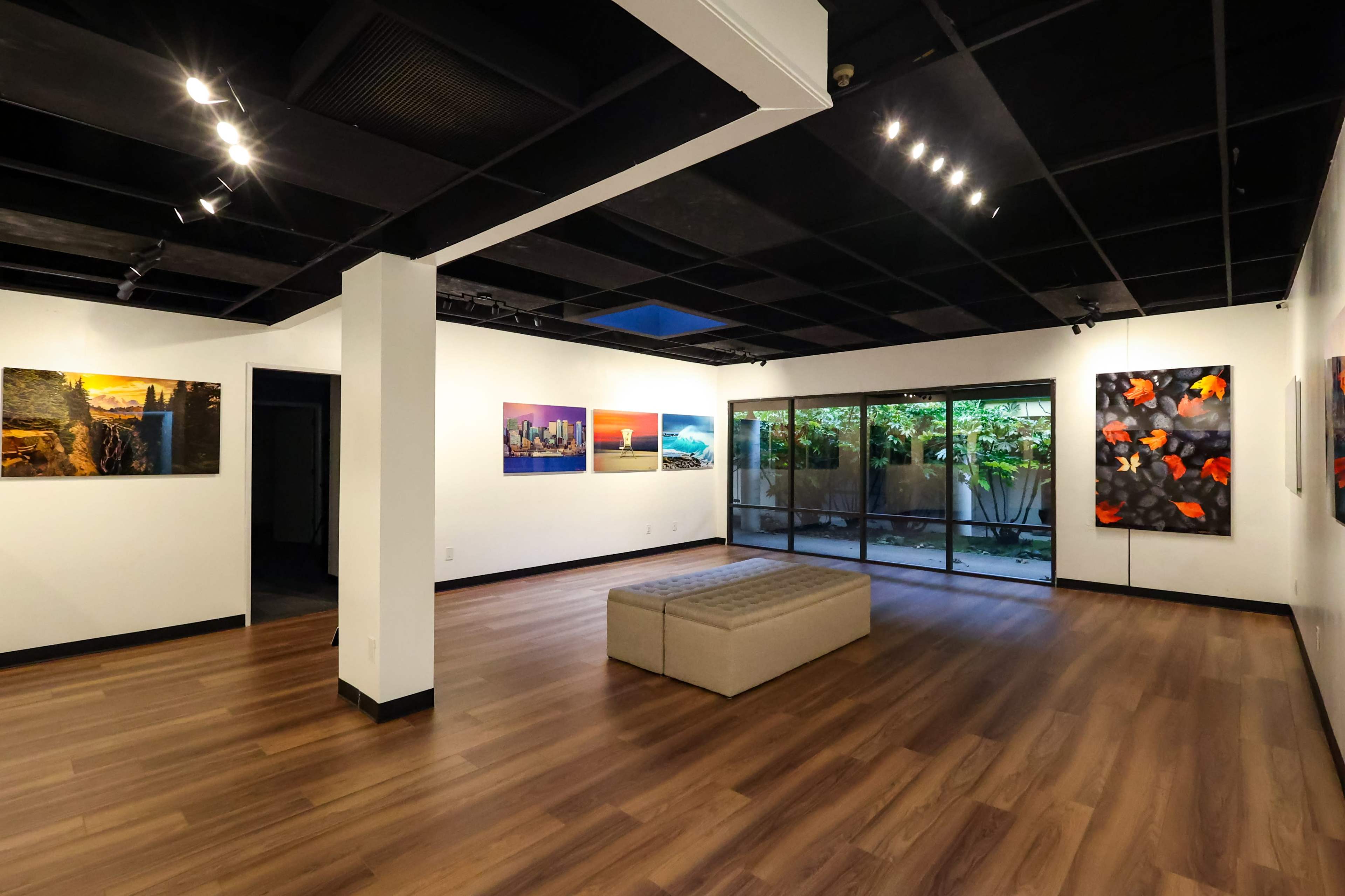 An art gallery interior with white walls, wooden flooring, and several framed artworks displayed along the walls, including a large glass window overlooking a garden.