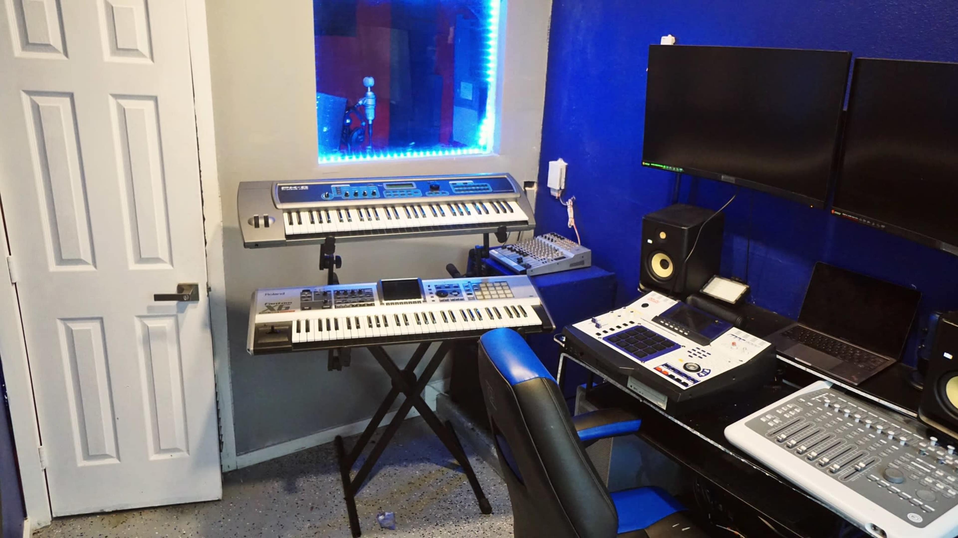 A music production setup in a room, featuring two keyboards, a computer station with multiple monitors, and a door.