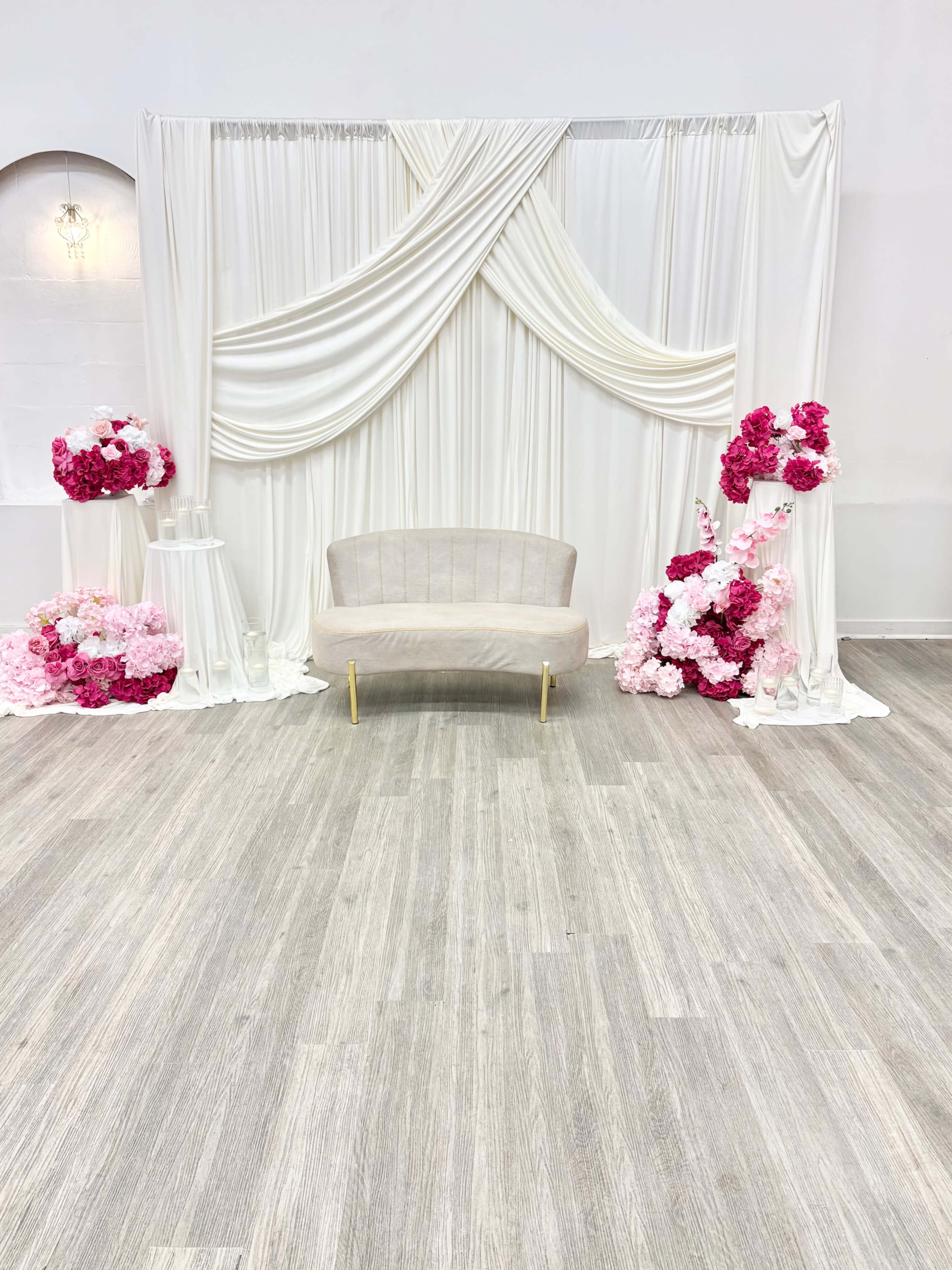 The image shows a simple event setup featuring a draped white backdrop with pink floral arrangements on either side and a light-colored sofa in the center.
