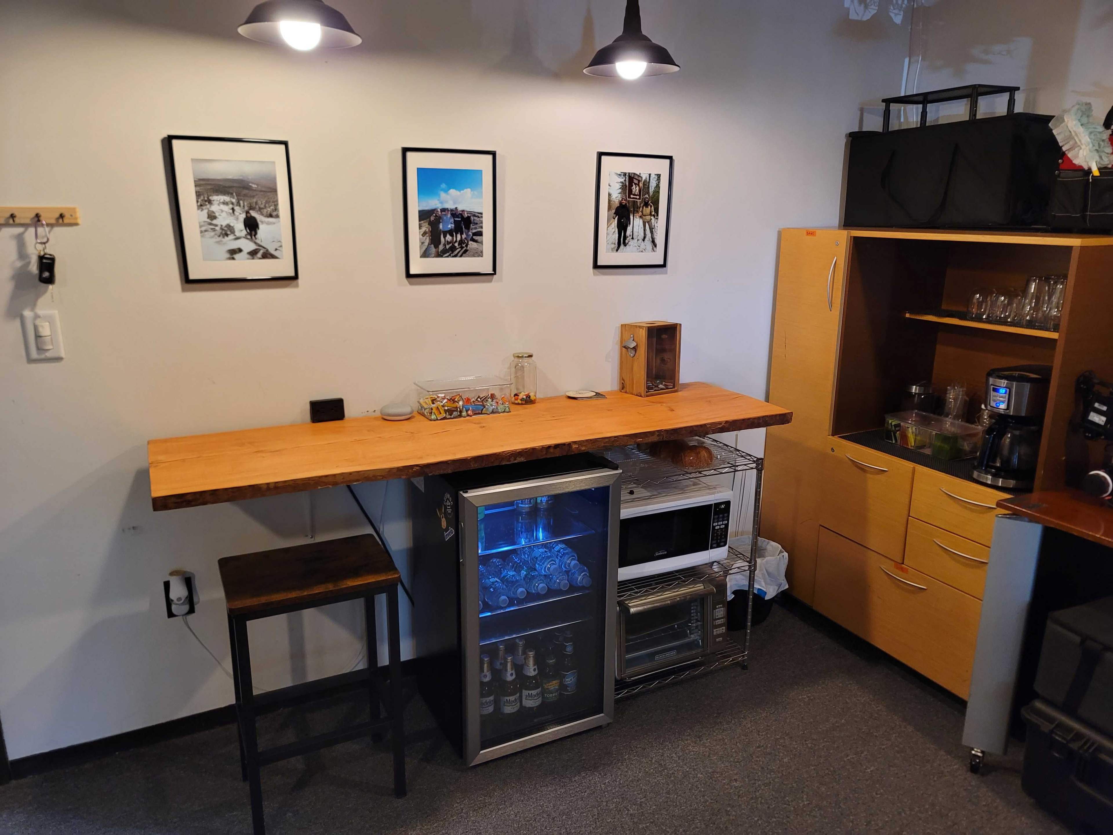 A small kitchen space with a wooden bar counter, framed pictures on the wall, and a glass-front refrigerator beneath the counter.