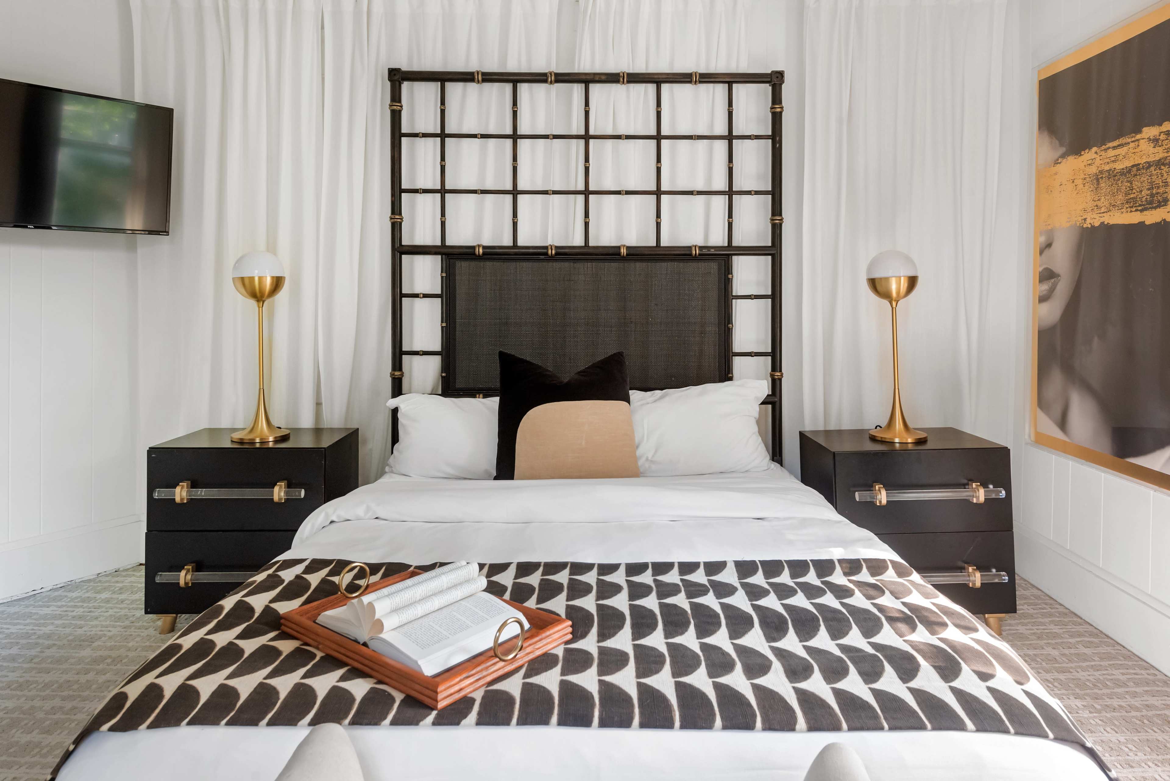 The image shows a modern bedroom featuring a bed with a dark headboard, two gold table lamps on nightstands, a decorative throw blanket, and a flat-screen TV mounted on the wall.