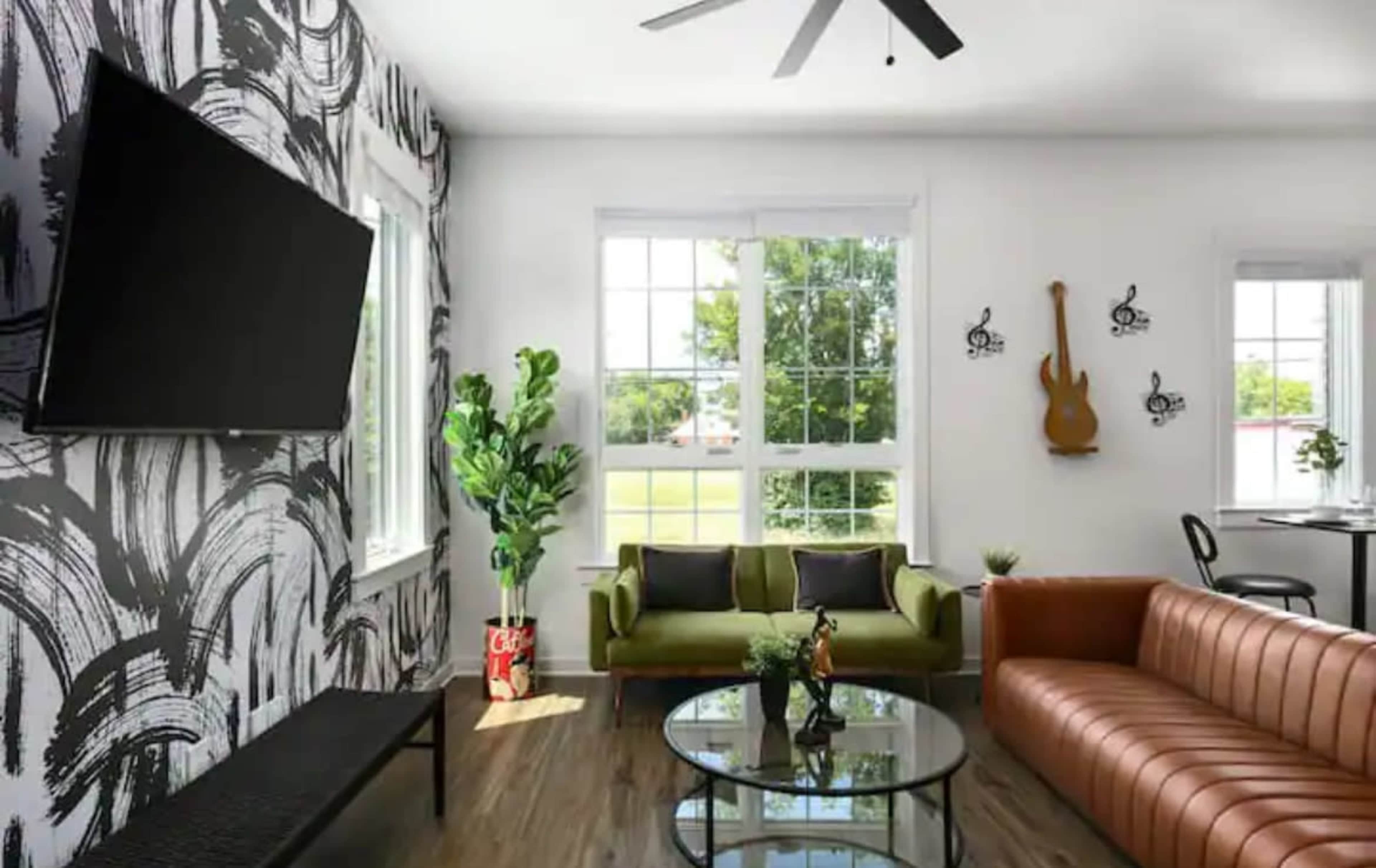 The room features a mix of modern and vintage furniture, including a brown leather sofa, a green couch, and a wall-mounted TV, all set against a decorative black and white wallpaper.
