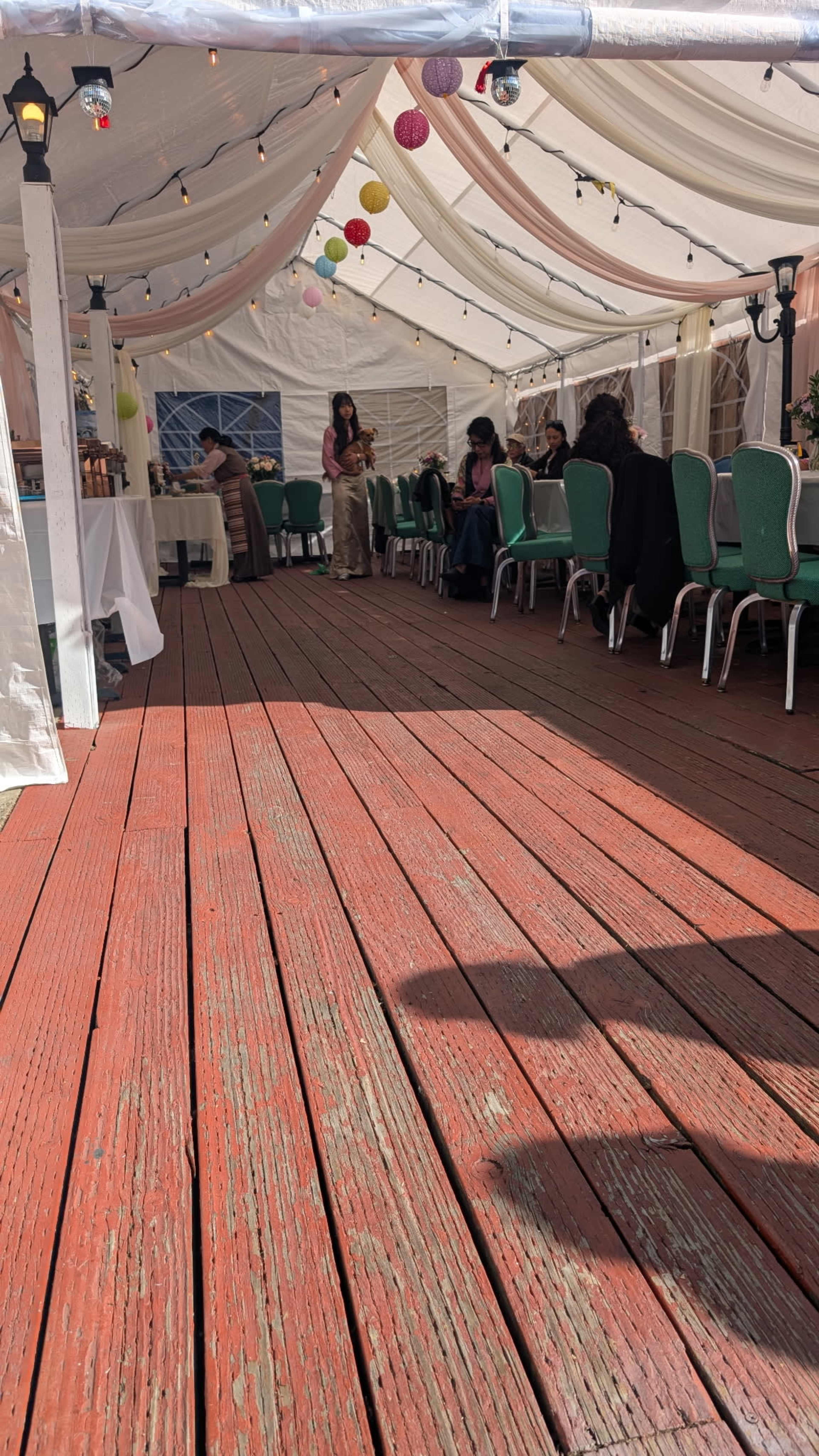 The image shows a partially filled outdoor tent with green chairs, wooden flooring, and decorative lights hanging overhead.