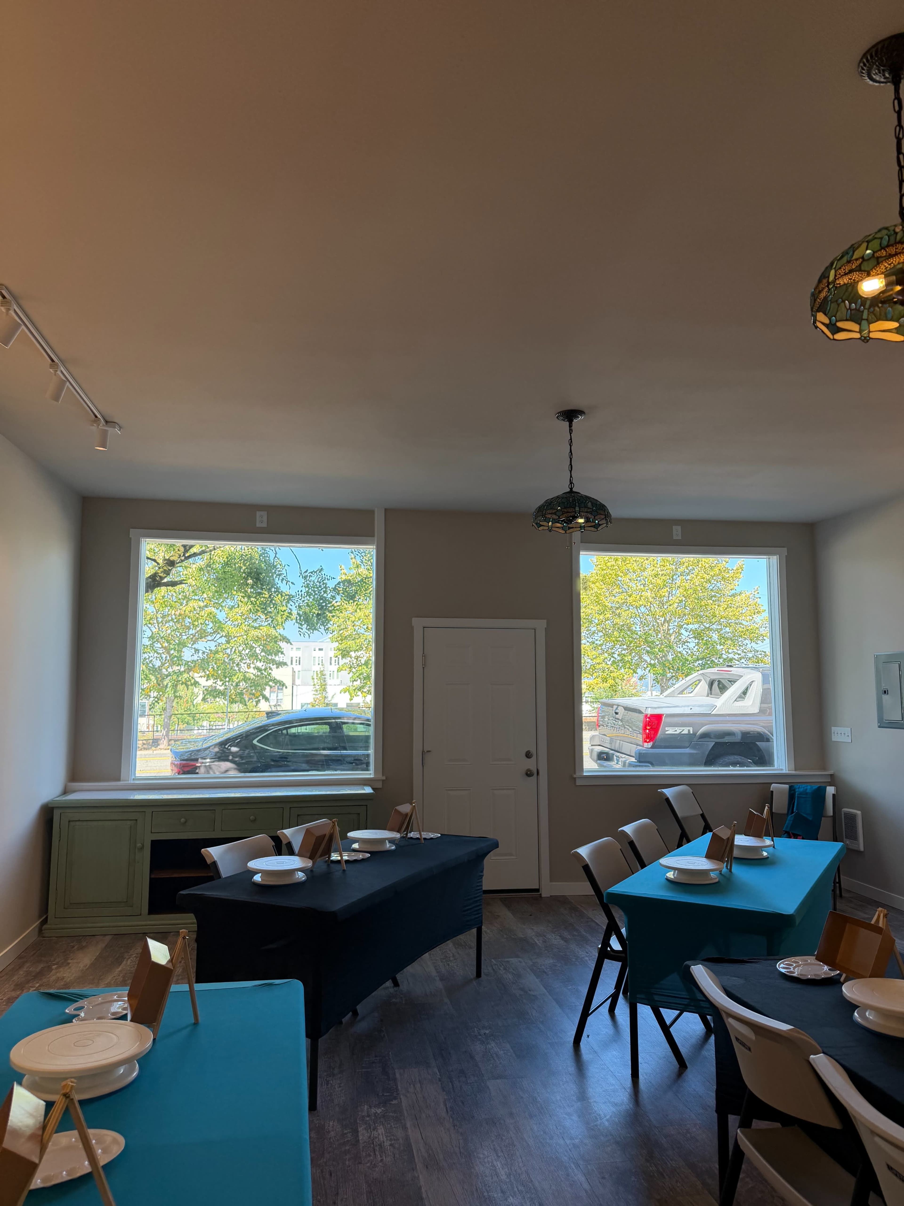 The image shows a dining area with two large windows, a black table set for a meal, and chairs arranged around it, featuring a sideboard against the wall.