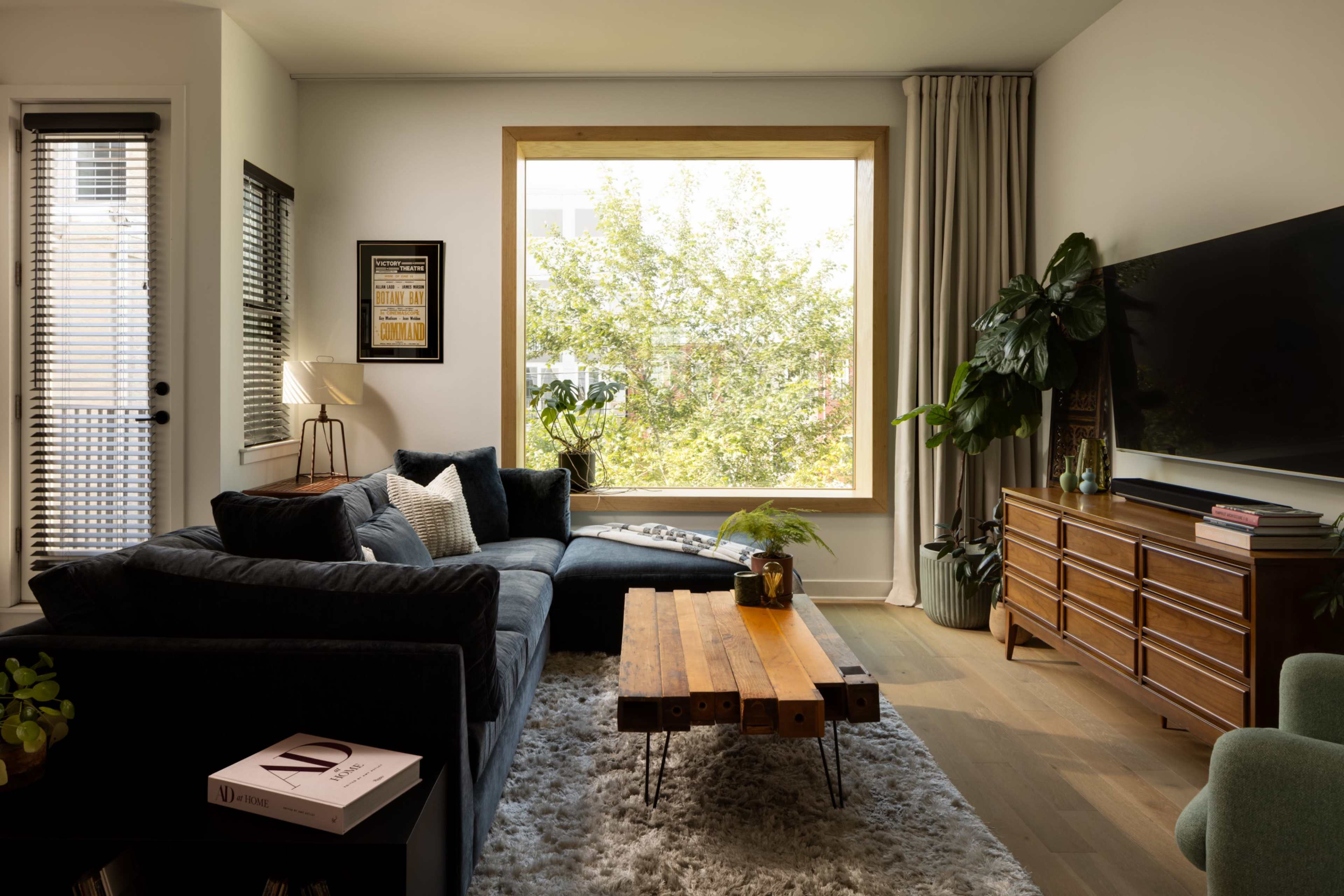A modern living room features a dark sectional sofa, a wooden coffee table, and a large window overlooking greenery.
