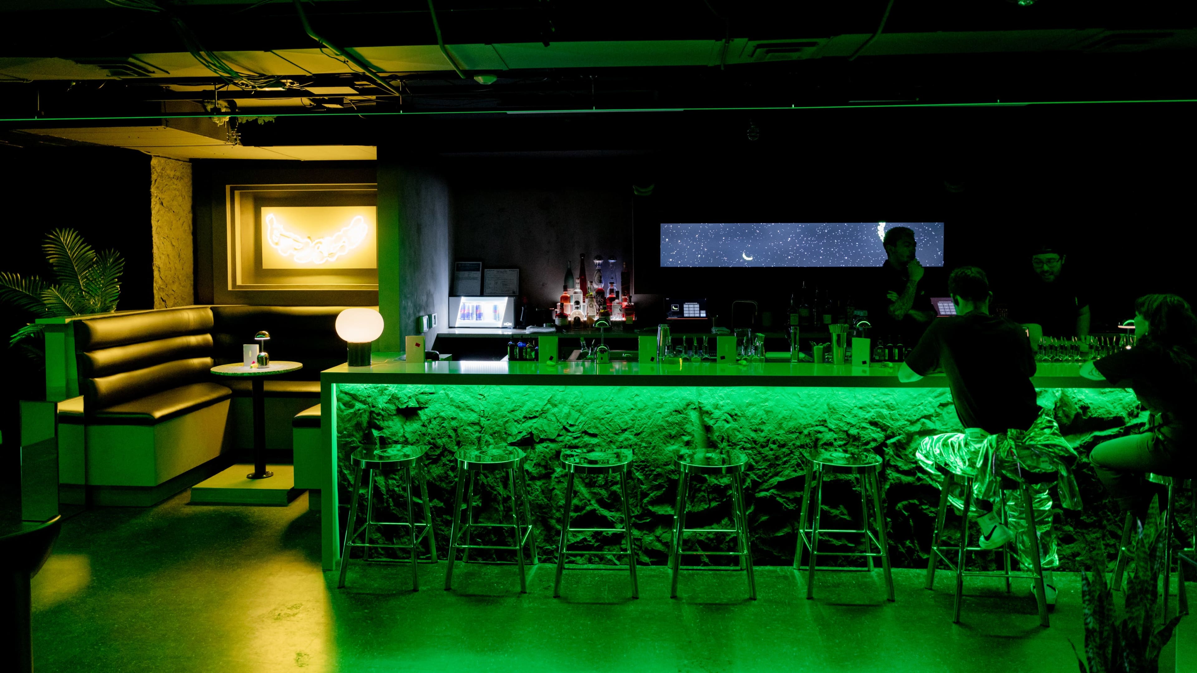 The image shows a modern bar with an illuminated green countertop and a dark interior featuring a booth and a large screen displaying stars.