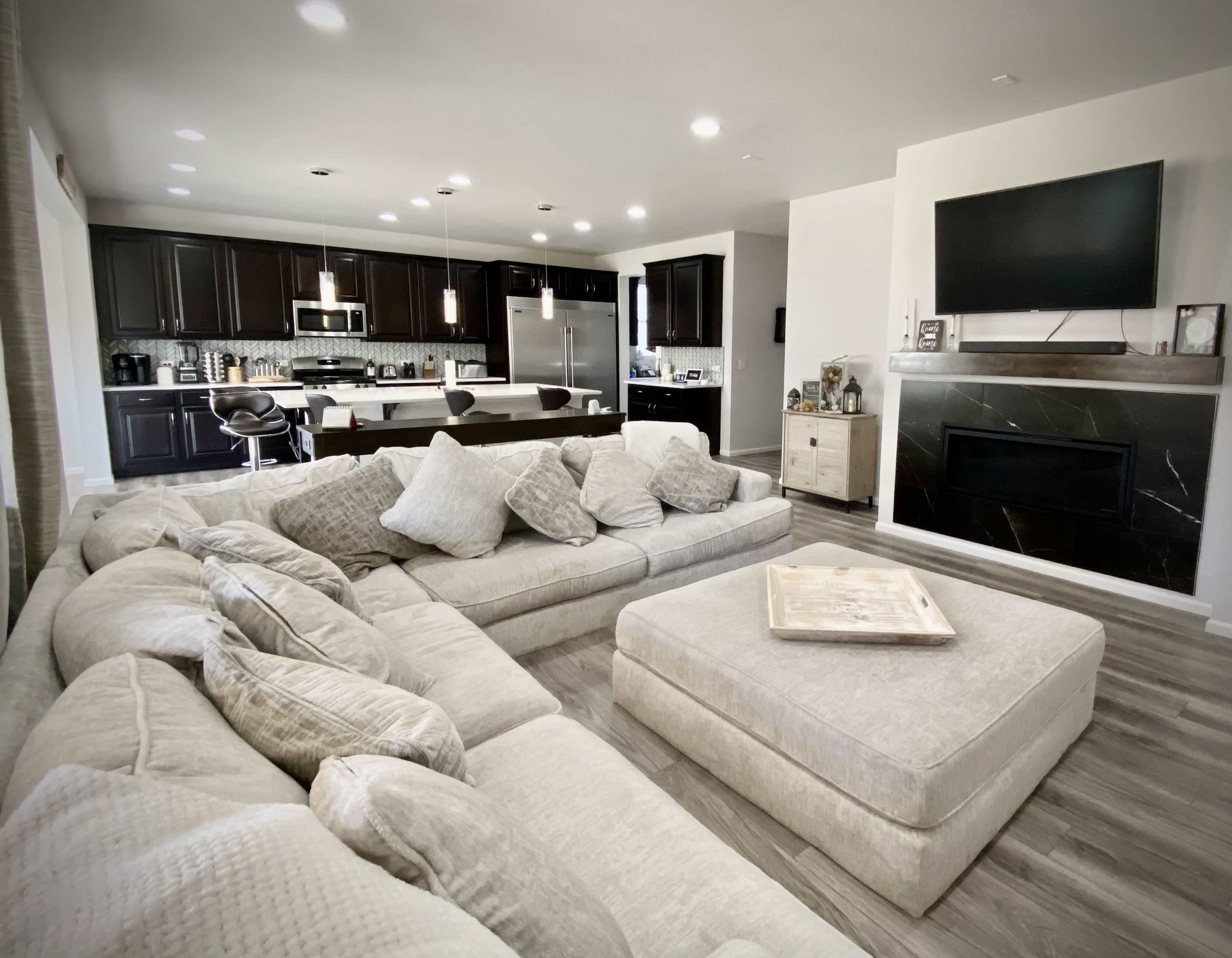 A modern living room featuring a large sectional sofa, an open kitchen with dark cabinetry, and a flat-screen TV mounted above a fireplace.
