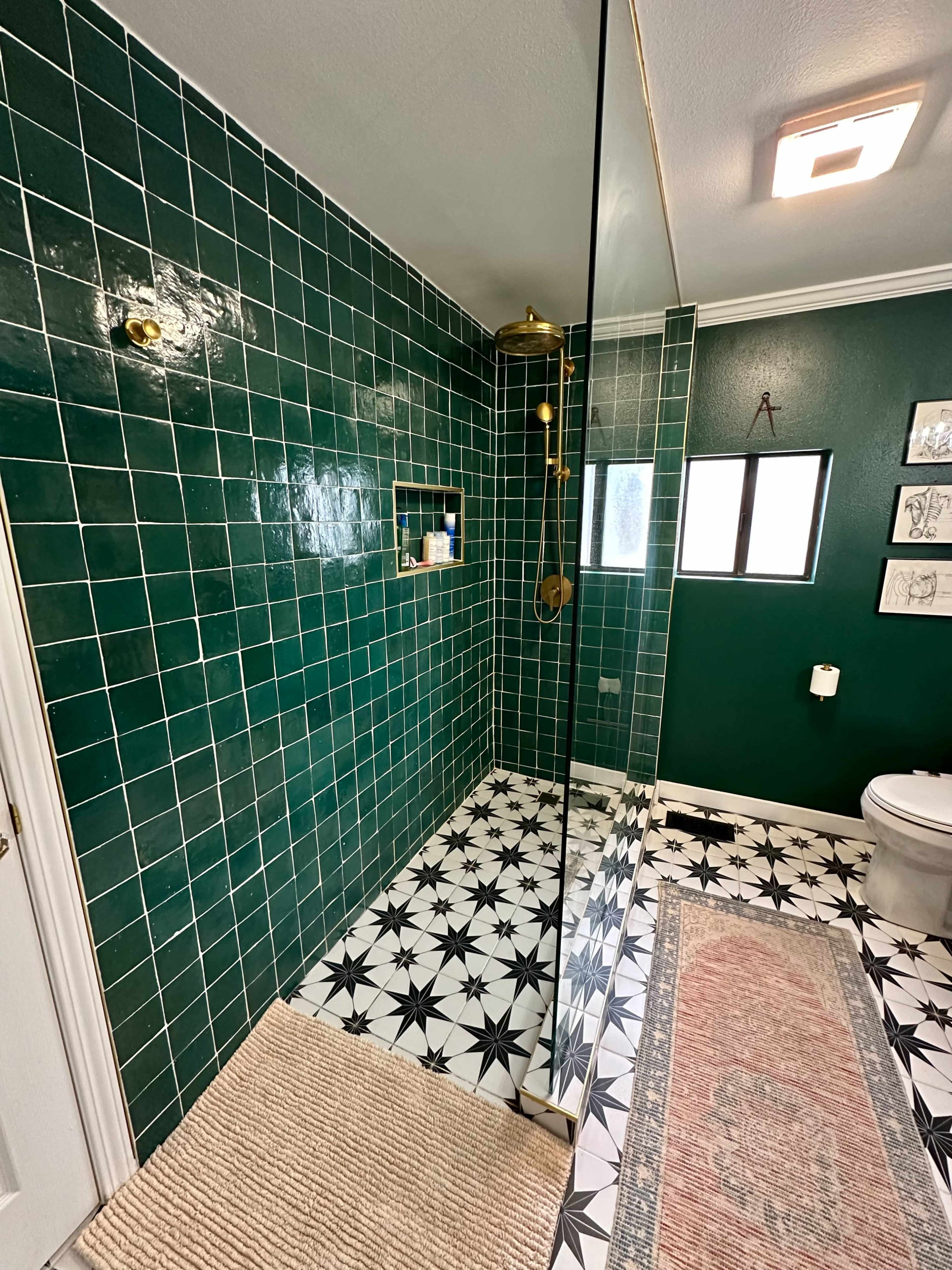 The image shows a modern bathroom featuring green tiled walls, a glass shower enclosure, a patterned black and white tiled floor, and a toilet beside a window.