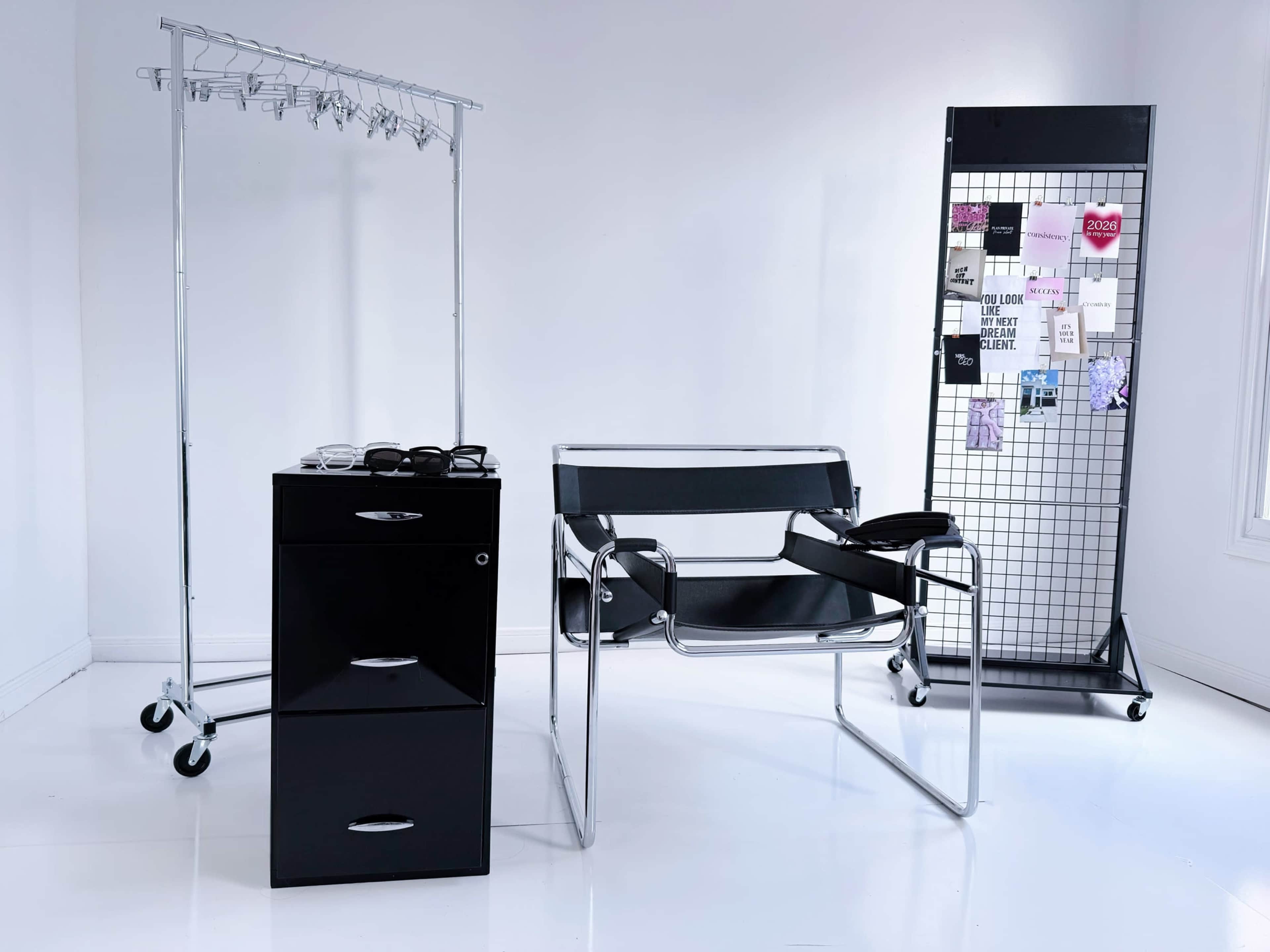 The image shows a minimalist interior featuring a black rolling storage unit, a modern chair, a clothing rack with hangers, and a grid display board with various notes and images.