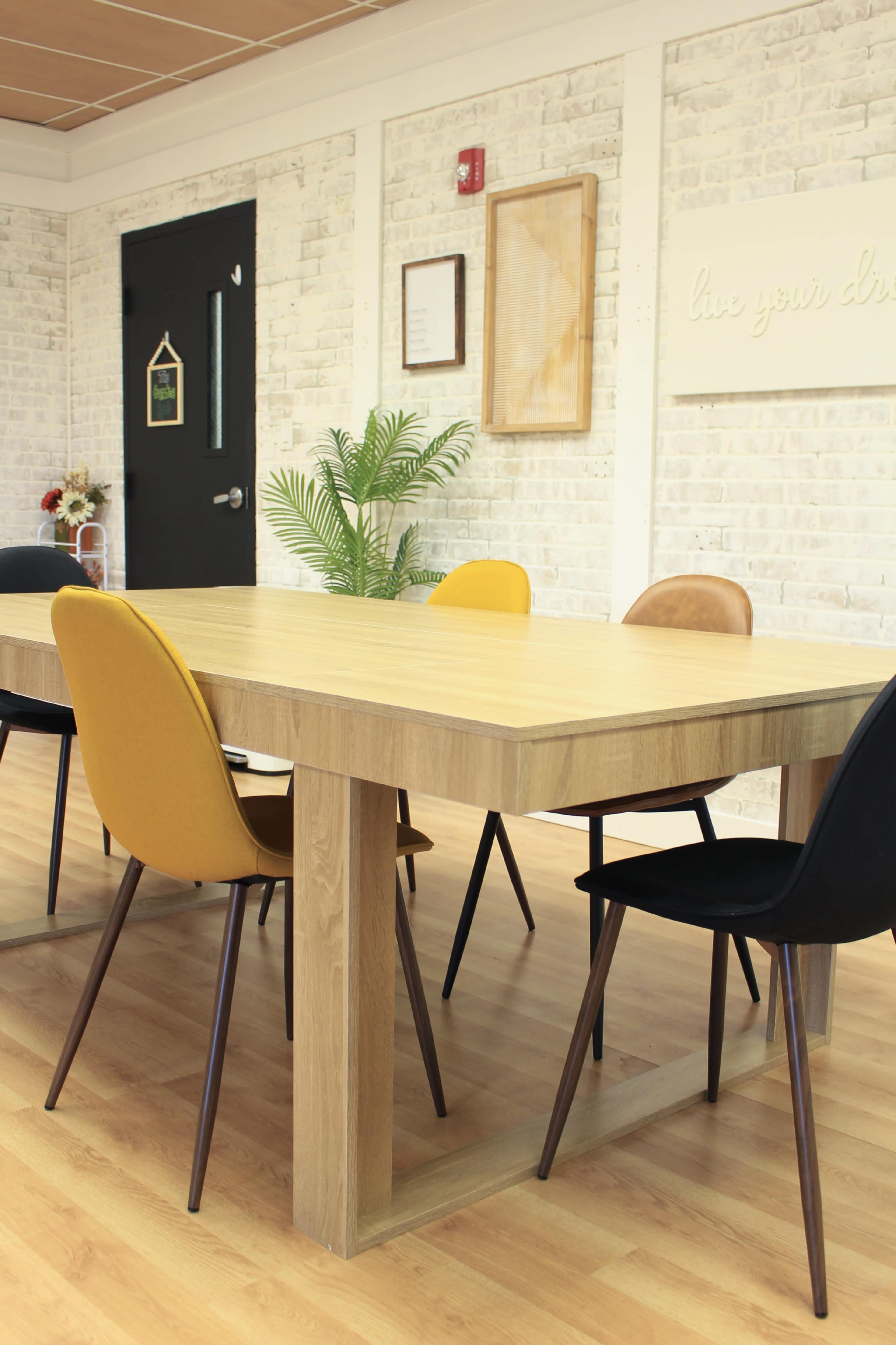 A wooden table with four chairs in various colors is set in a room with white brick walls and minimal decor.