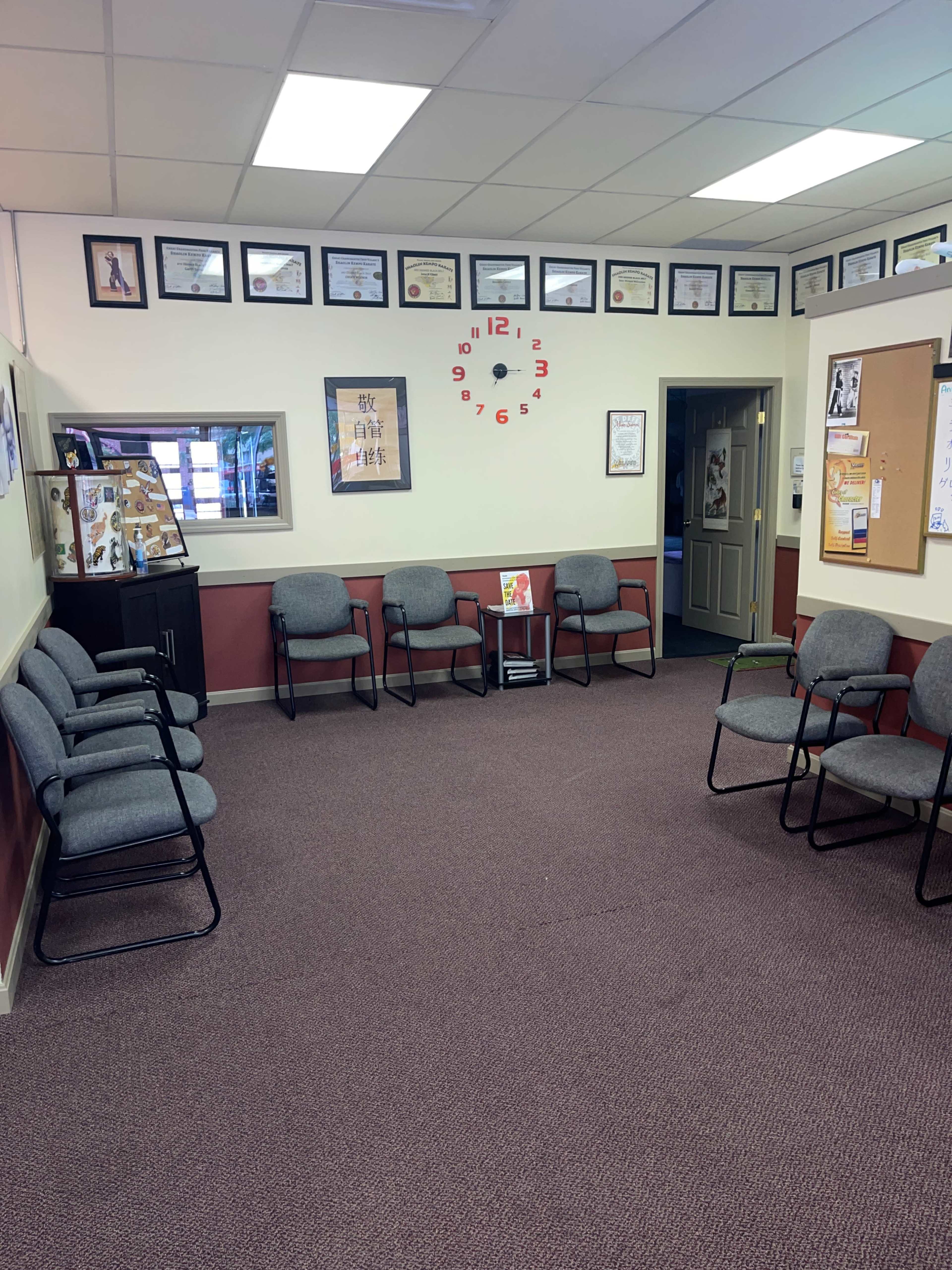 A waiting area with several gray chairs arranged around a central space, featuring framed certificates and a clock on the wall.