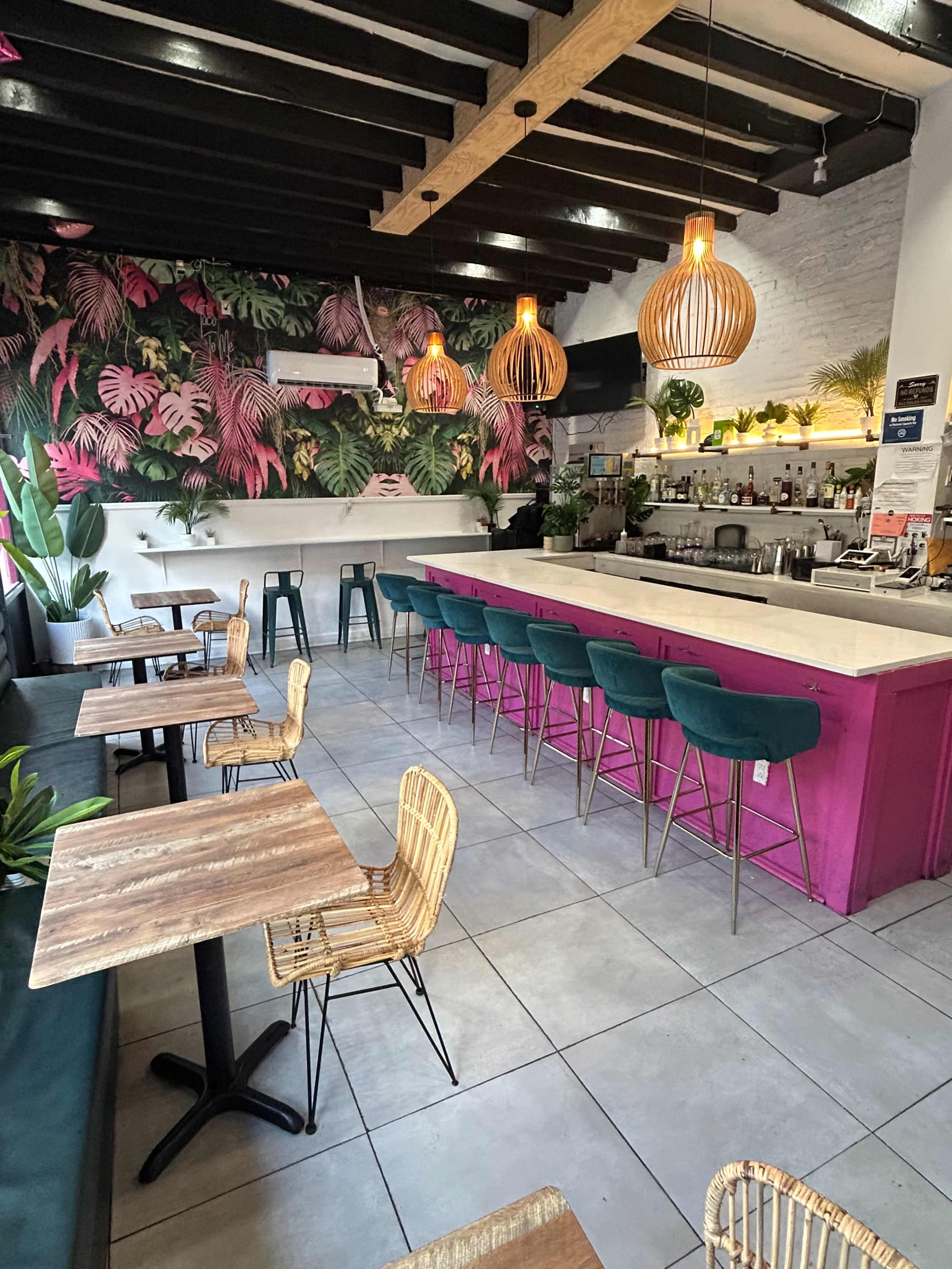 The image shows a brightly decorated café with a pink bar, tropical wallpaper, wooden tables, and rattan chairs arranged around the seating area.
