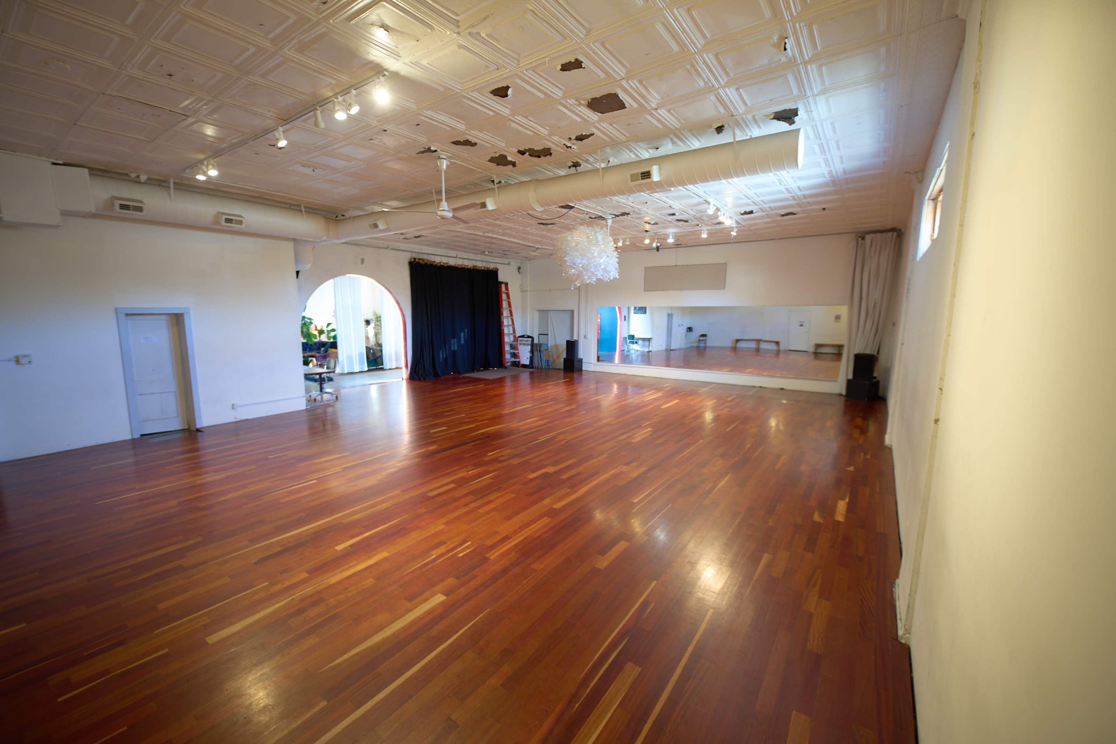 The image shows a spacious dance studio with polished wooden floors, a mirrored wall, and various lighting fixtures.