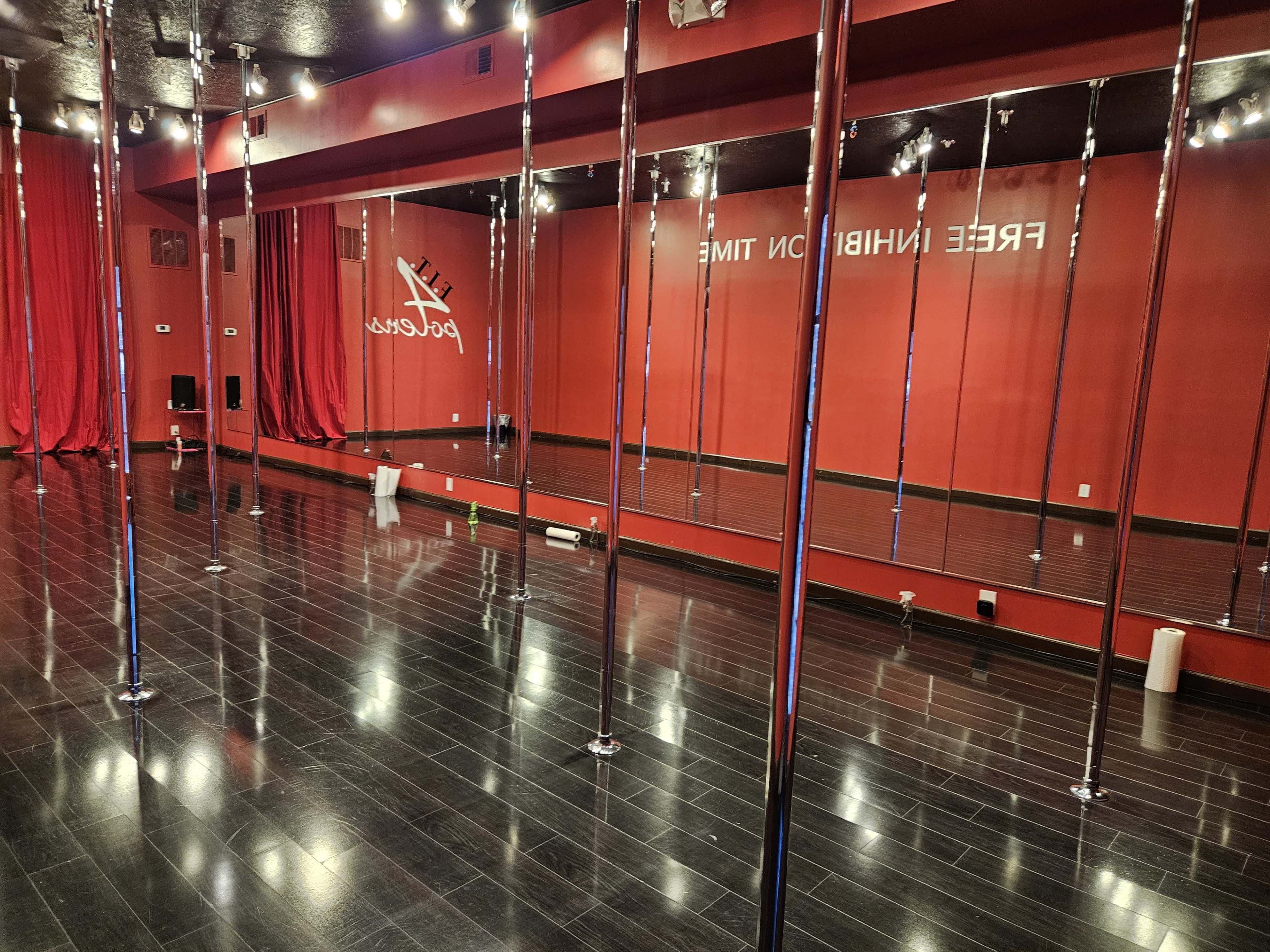 A dance studio with polished black floors, multiple chrome poles, and red curtains on the walls.