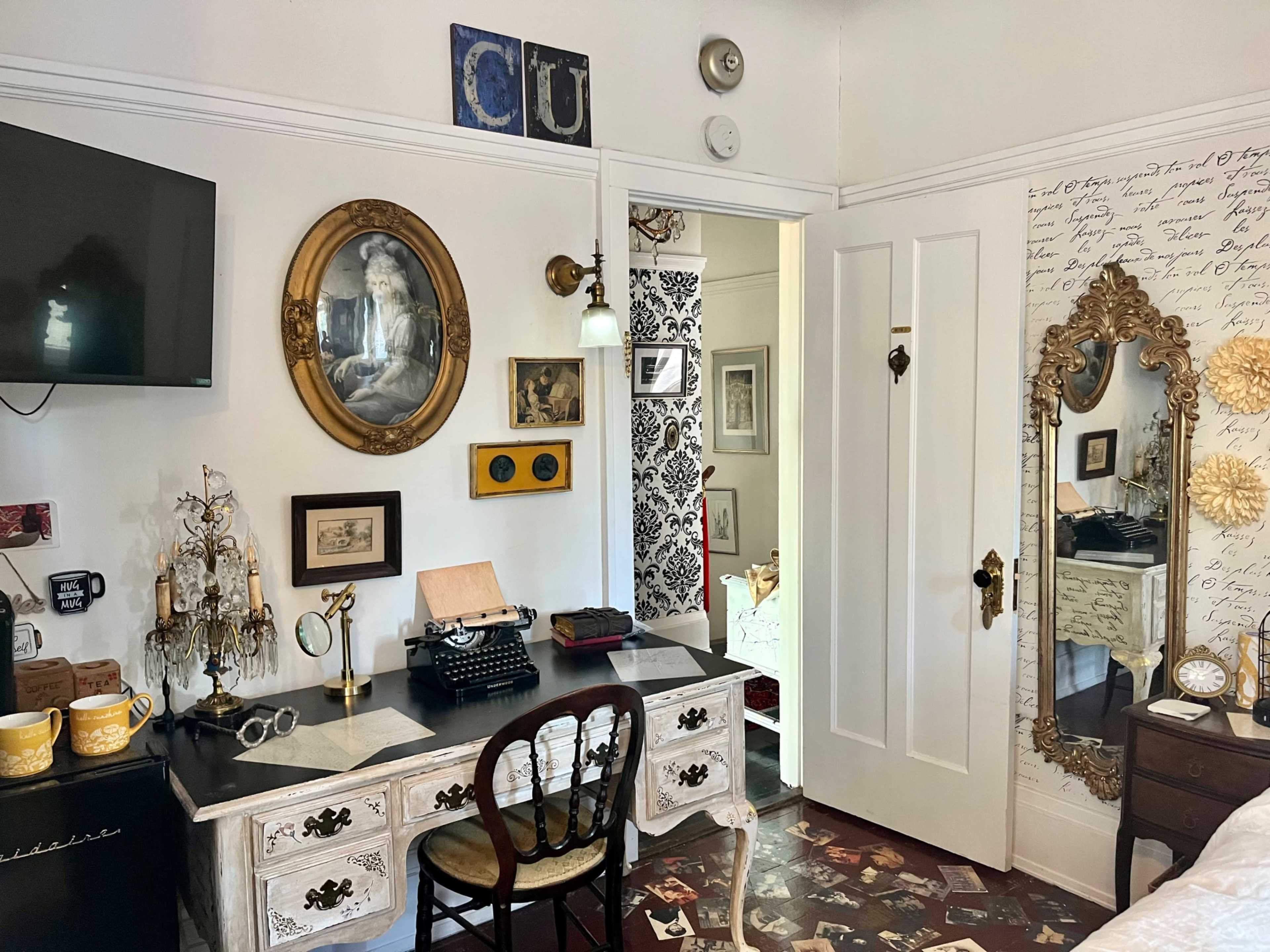 The scene depicts a cozy home office featuring a vintage typewriter on a black and white desk, framed artwork on the walls, and a doorway leading to another room.