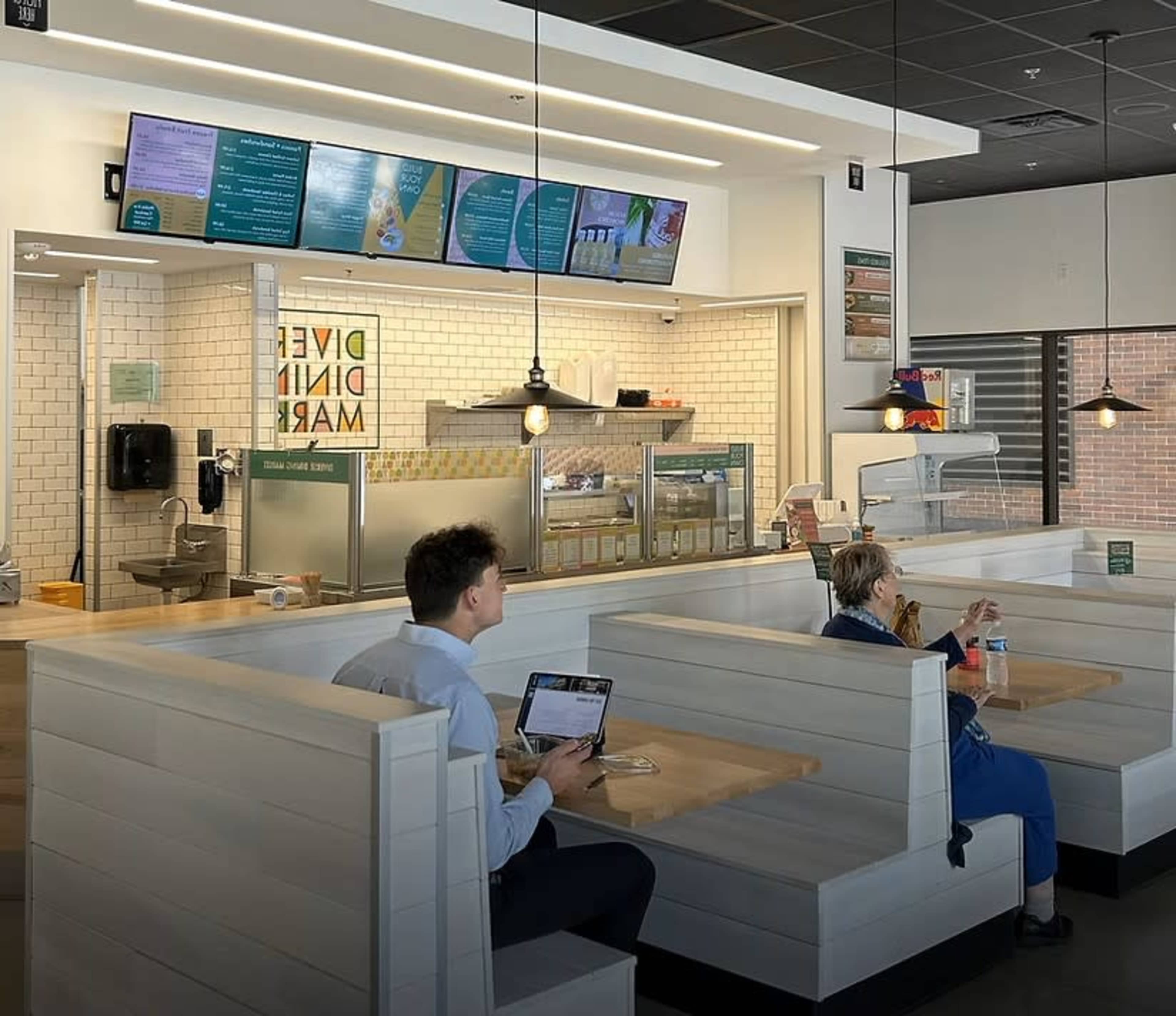 A spacious, modern café features white wooden booths and a menu display above the ordering counter, with two patrons seated inside.