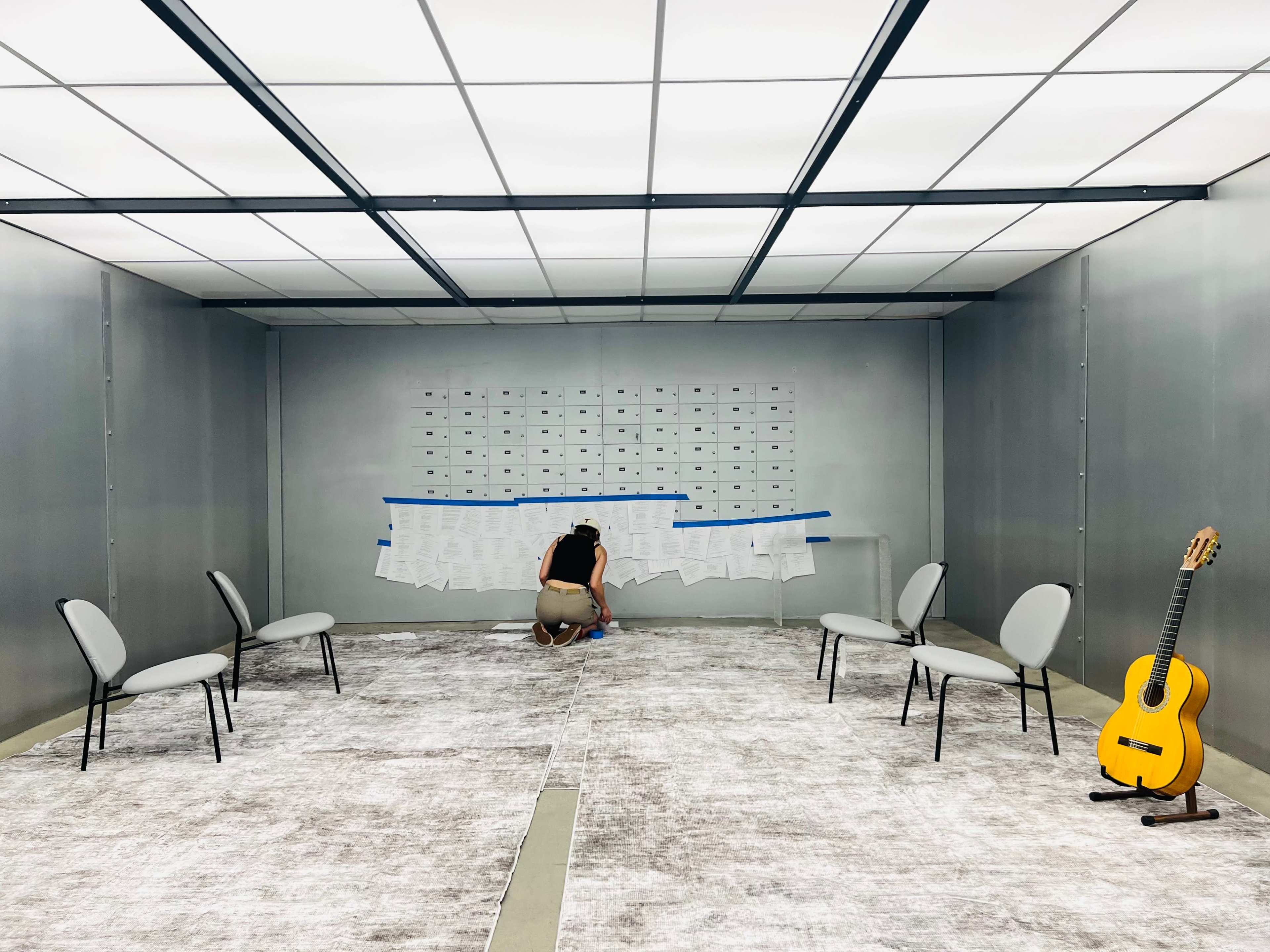 A person is kneeling in a sparsely furnished room with gray walls, placing paper on a wall, while a guitar rests on the floor beside empty chairs.