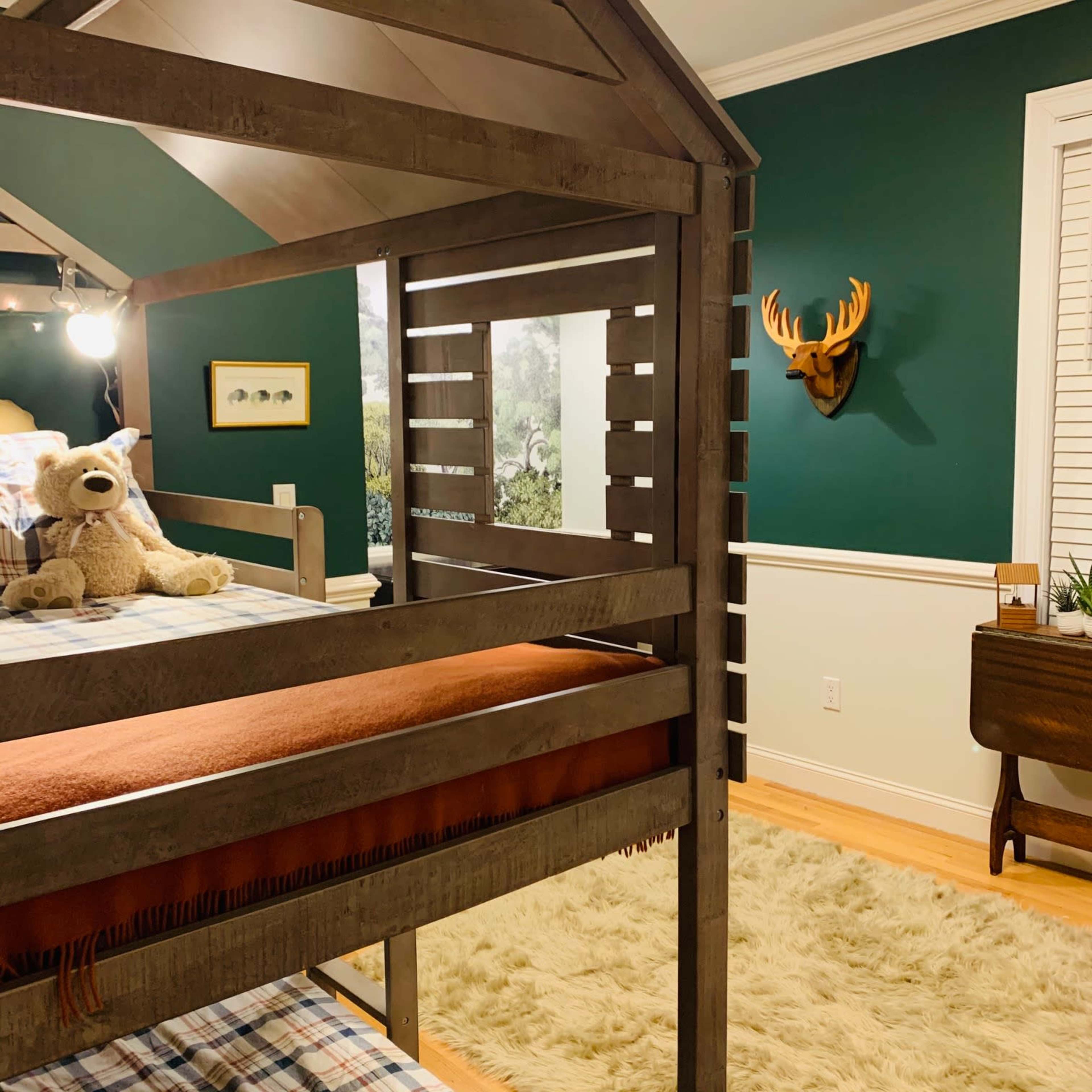 A wooden loft bed with a tent-like design is positioned in a room featuring green walls and decorated with a stuffed bear and deer head mount.