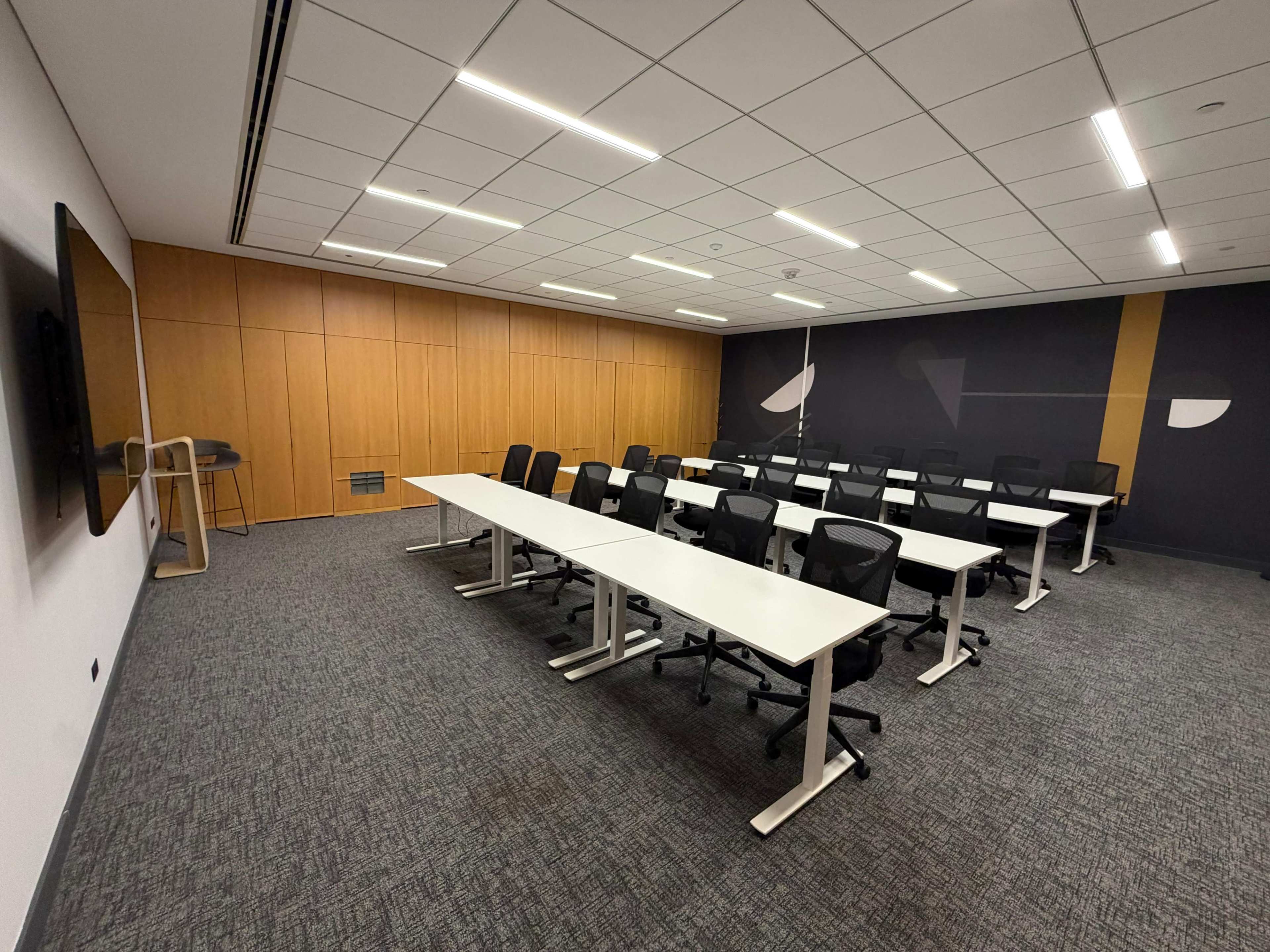 The image shows a well-lit meeting room with a series of white tables arranged in rows, black chairs, a wall-mounted screen, and wooden paneling.