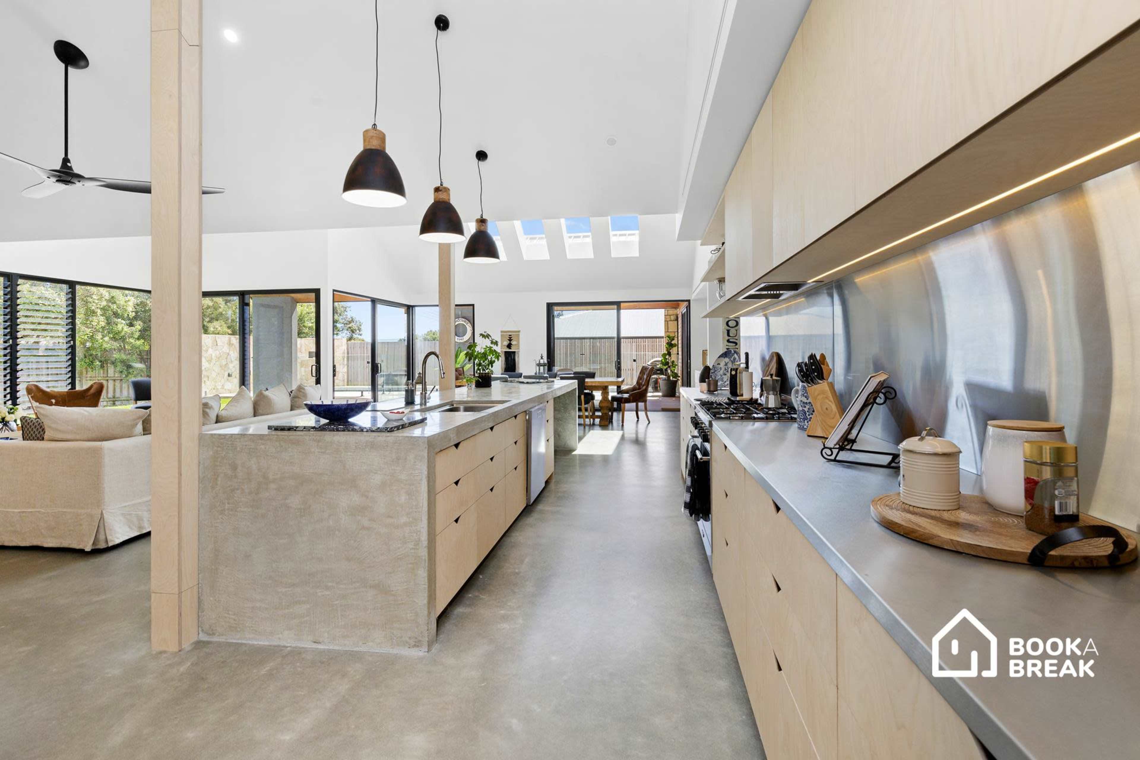 A modern kitchen with a long island countertop, wooden cabinetry, and large windows allowing natural light to fill the space.