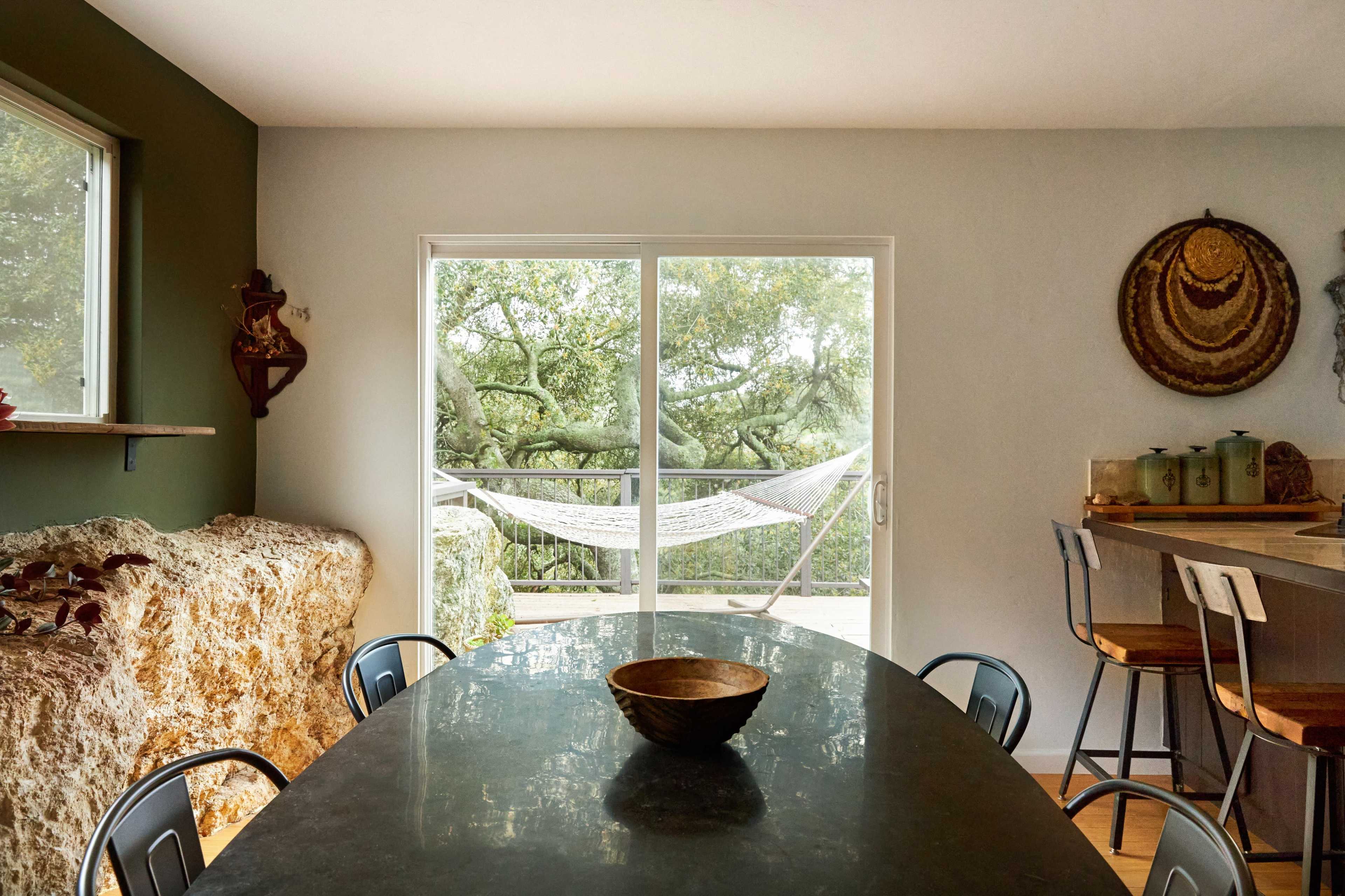 A dining area features a dark table with a wooden bowl, surrounded by chairs, and large windows revealing a view of a hammock outside.