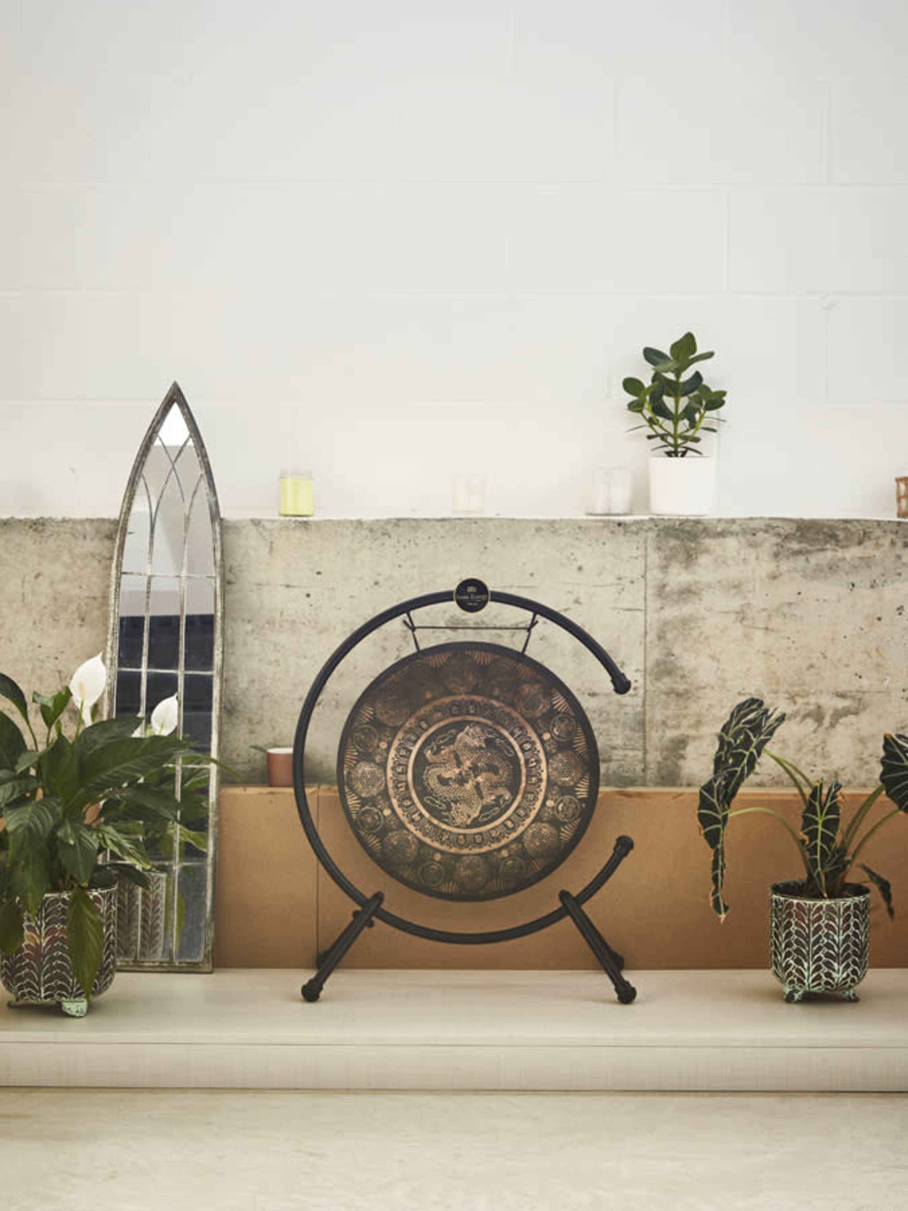 A decorative gong stands on a black metal frame against a concrete wall, surrounded by potted plants and a tall mirror.