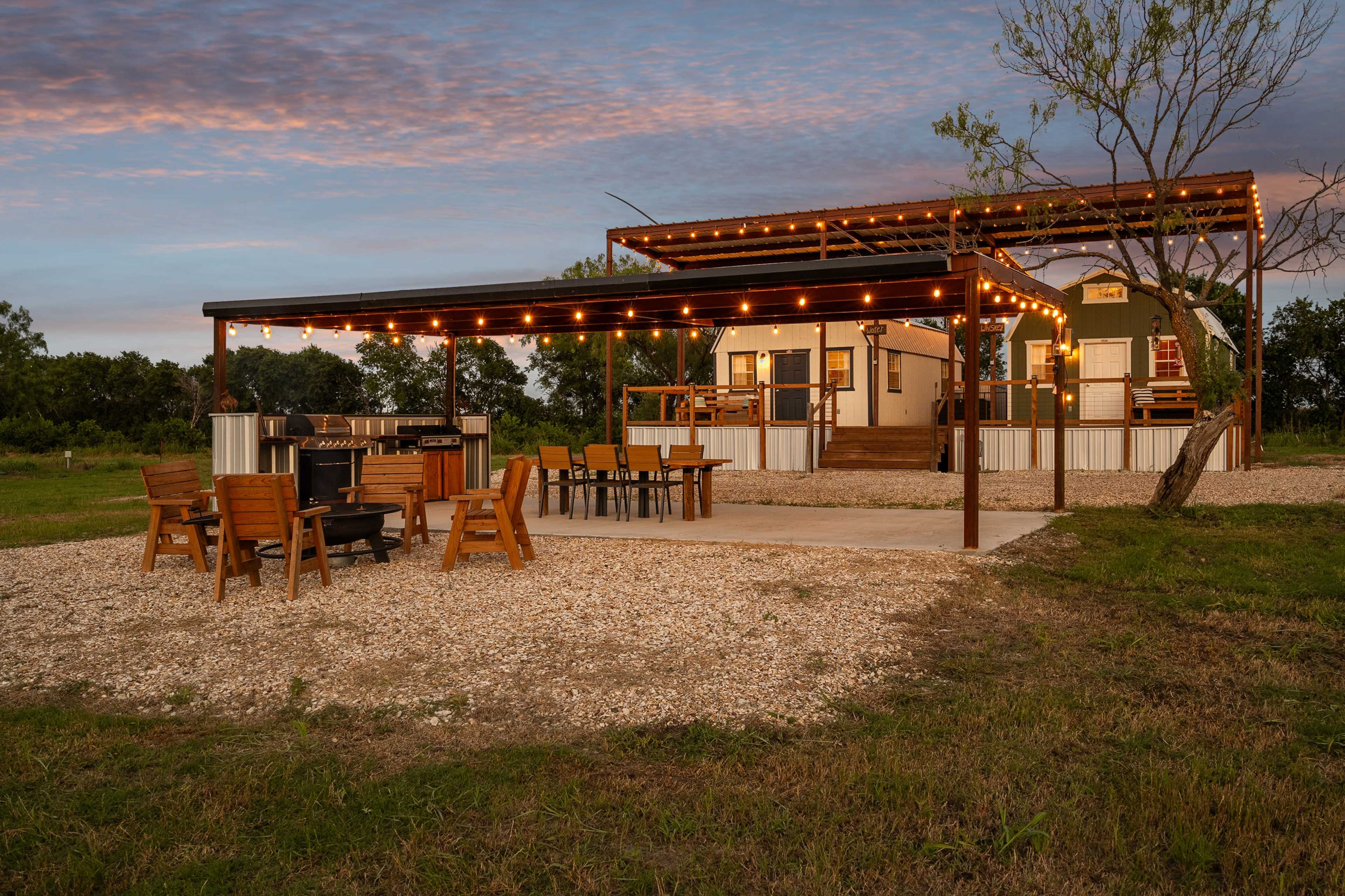 A modern cabin with a covered patio adorned with string lights is set within a grassy area, featuring outdoor seating and dining arrangements.