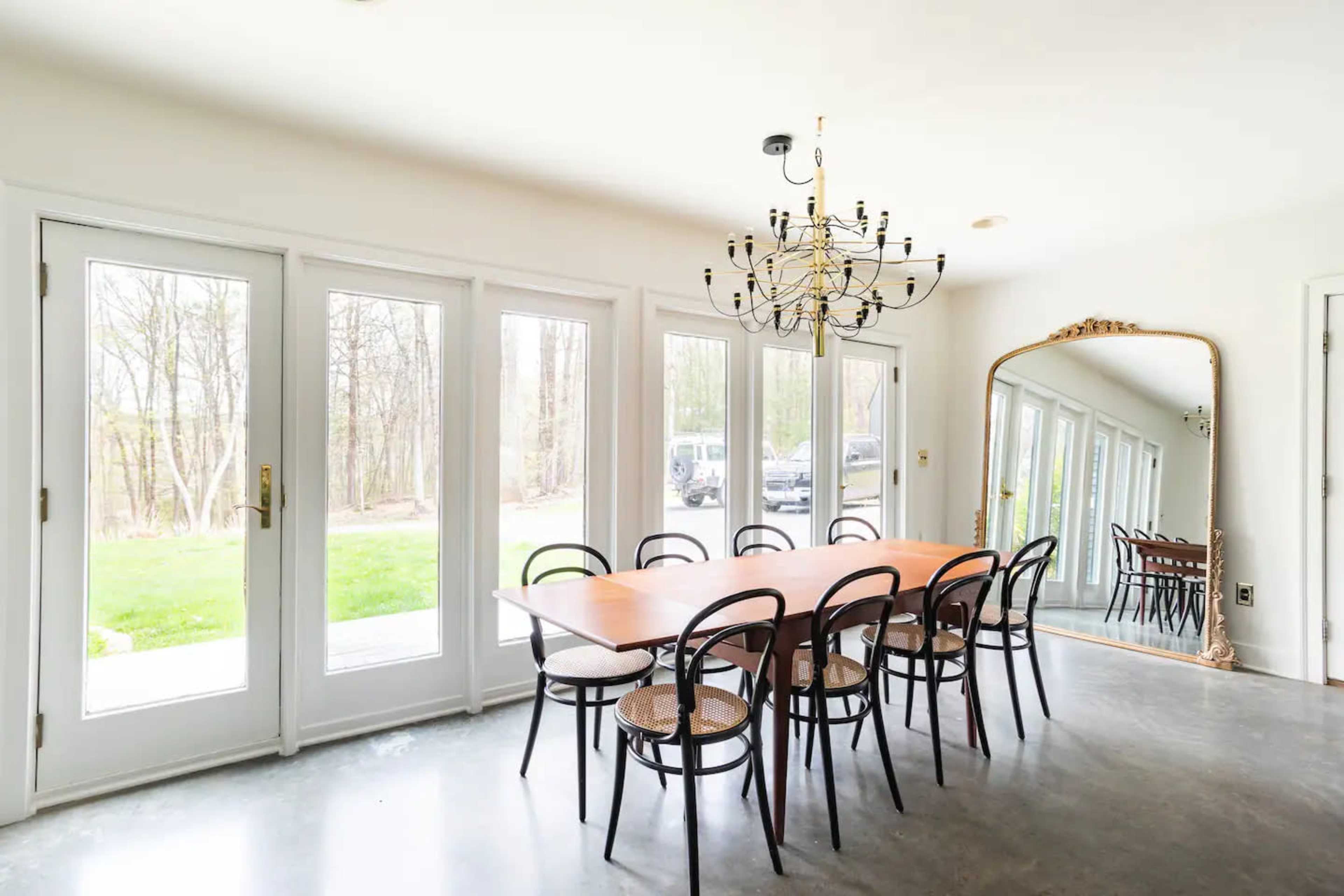 A dining area features a long wooden table with eight chairs, a chandelier overhead, large glass doors leading to an outdoor view, and a large decorative mirror on the wall.