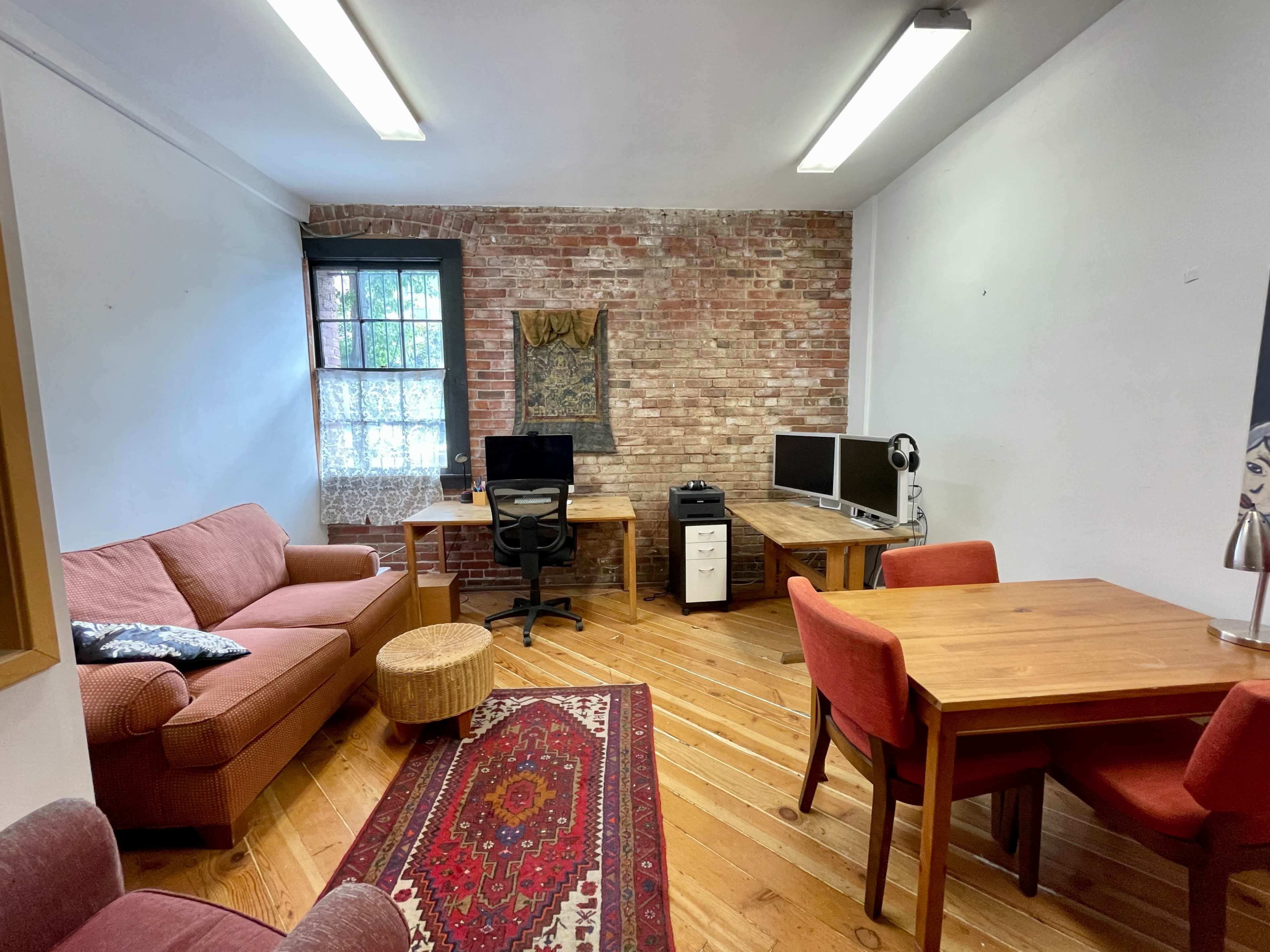 The image shows a cozy office space featuring a couch, two wooden desks with computers, a dining table with chairs, and exposed brick walls.