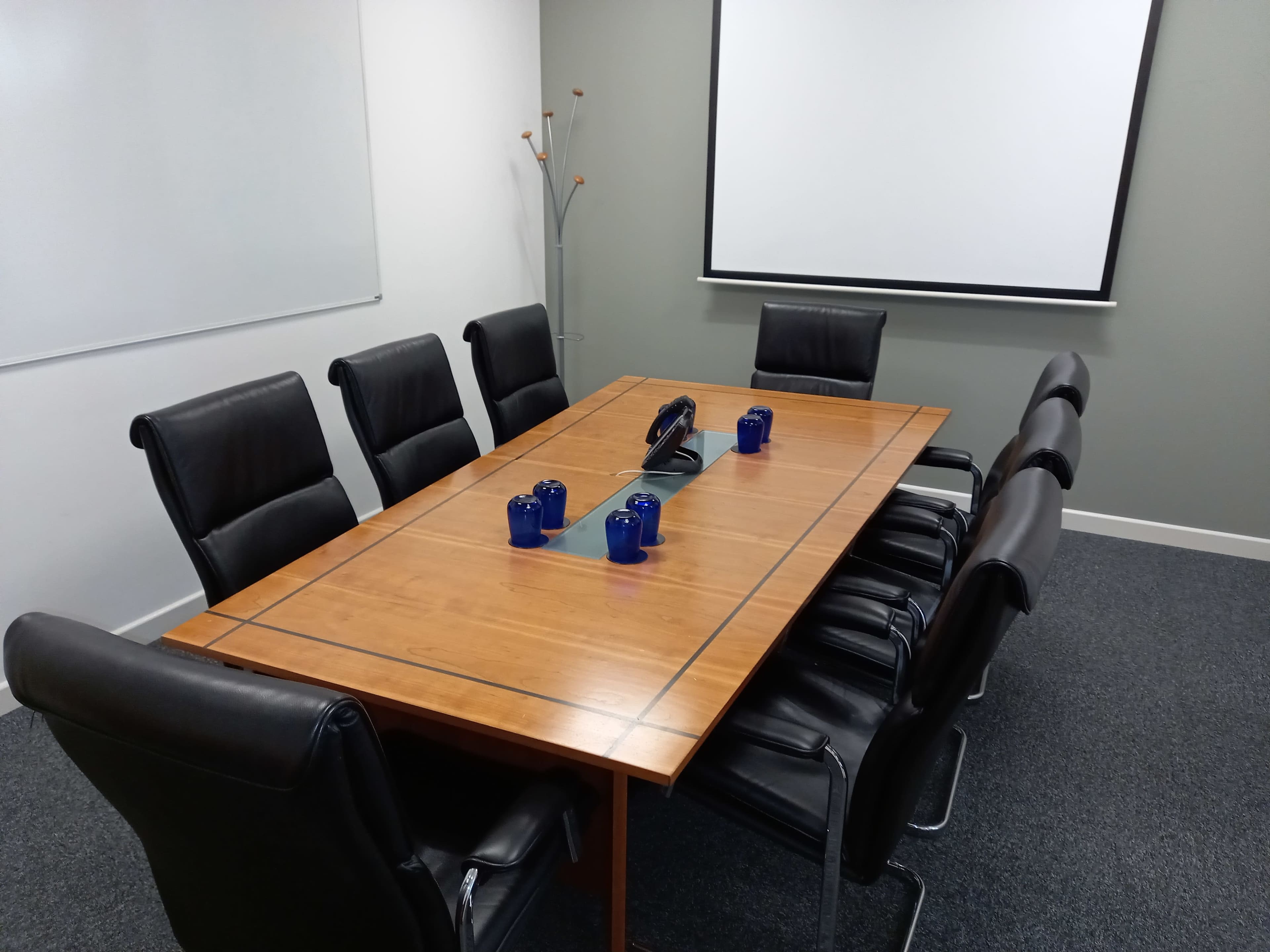 A conference room features a large wooden table surrounded by black leather chairs, with a projector screen in the front and blue mugs arranged in the center.