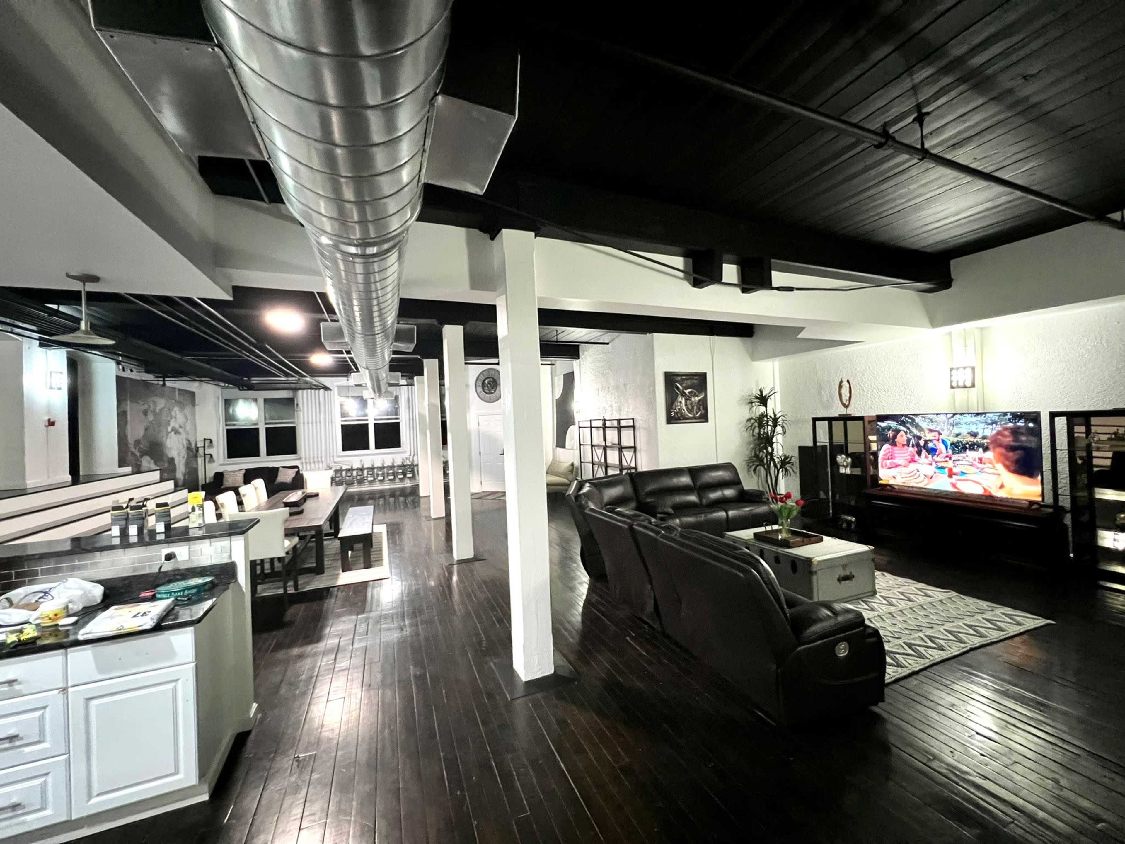 A spacious, modern living area with a black leather couch, a television displaying a colorful scene, and an open kitchen area with white cabinets.