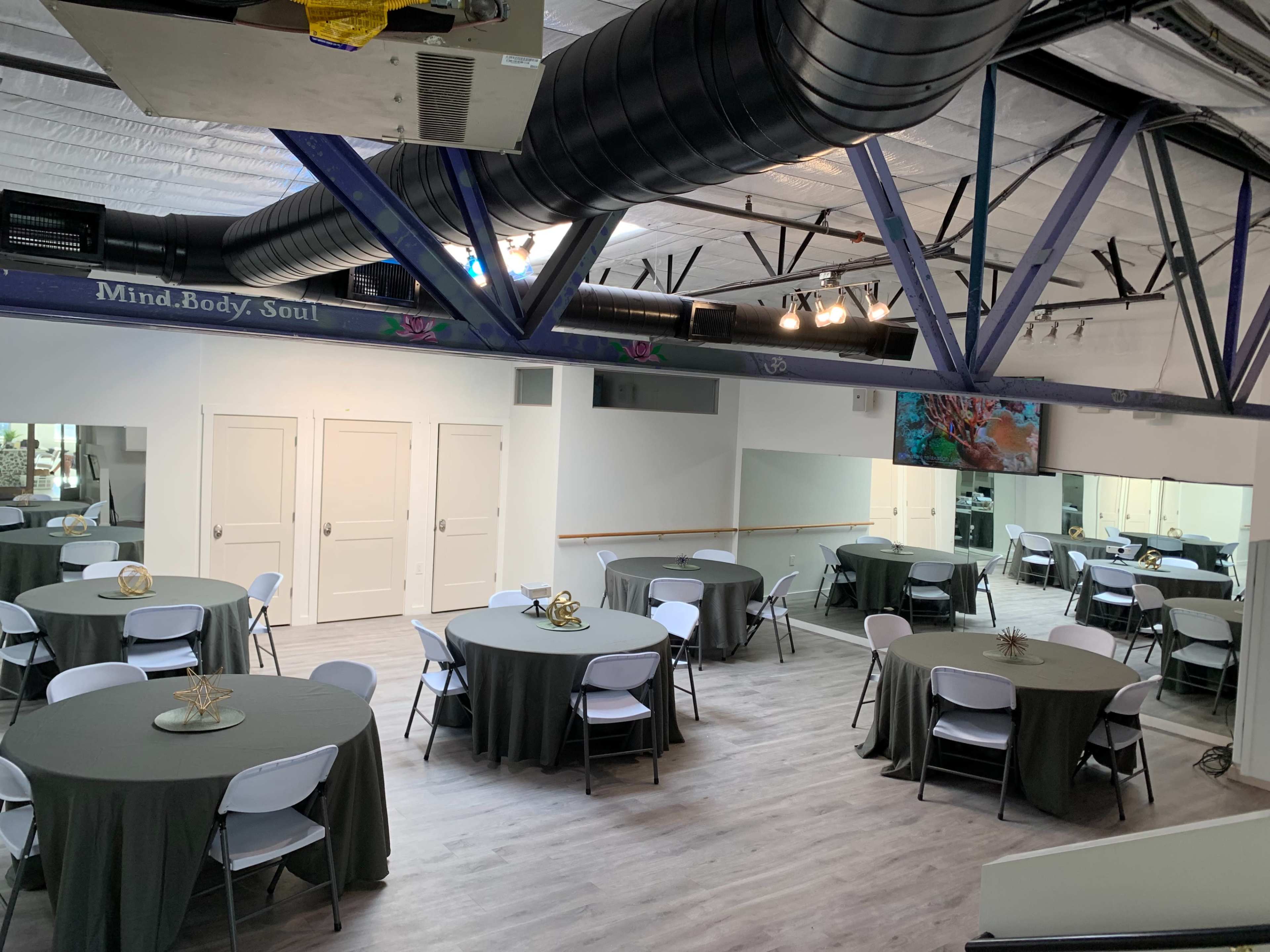 A spacious room with several round tables covered in gray tablecloths, set up for an event, and featuring large mirrors and a mounted screen on the wall.