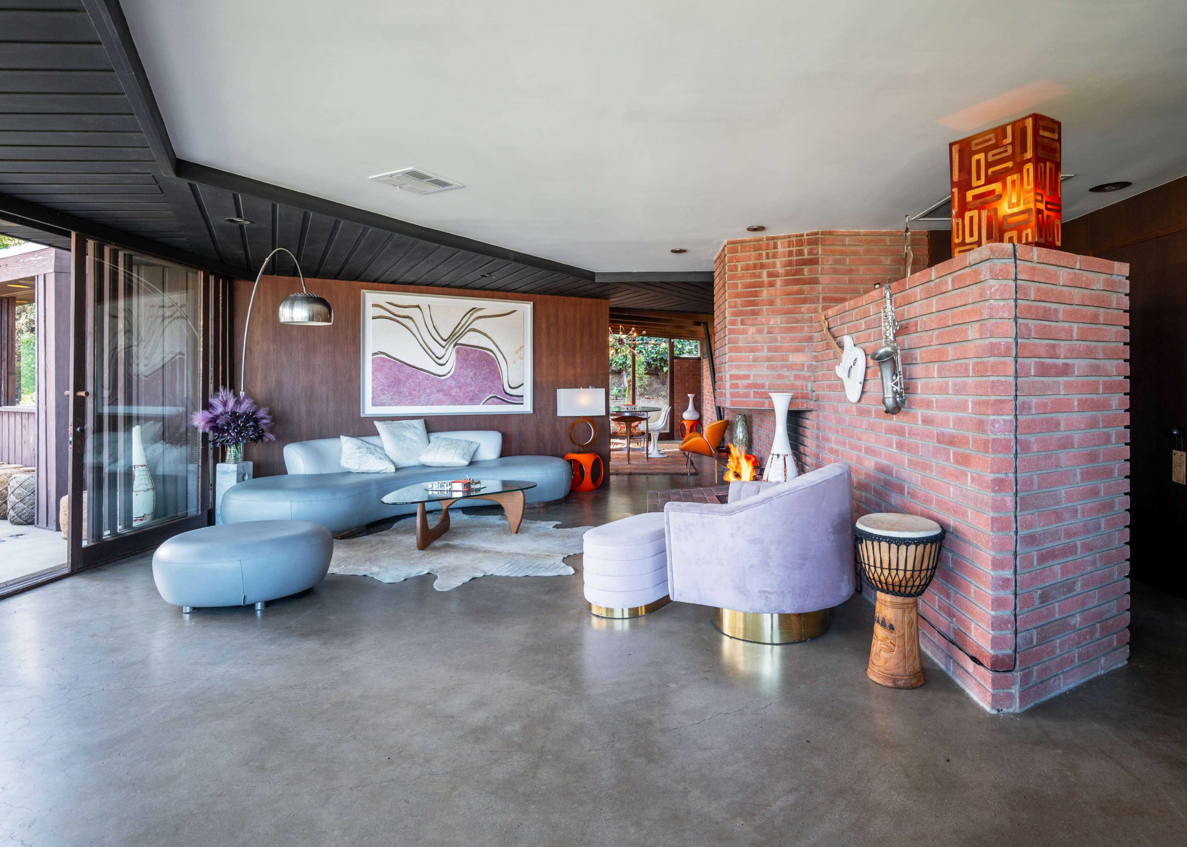 The image shows a modern living room featuring a turquoise sofa, a purple armchair, a glass coffee table, and a brick fireplace, with large windows providing natural light.