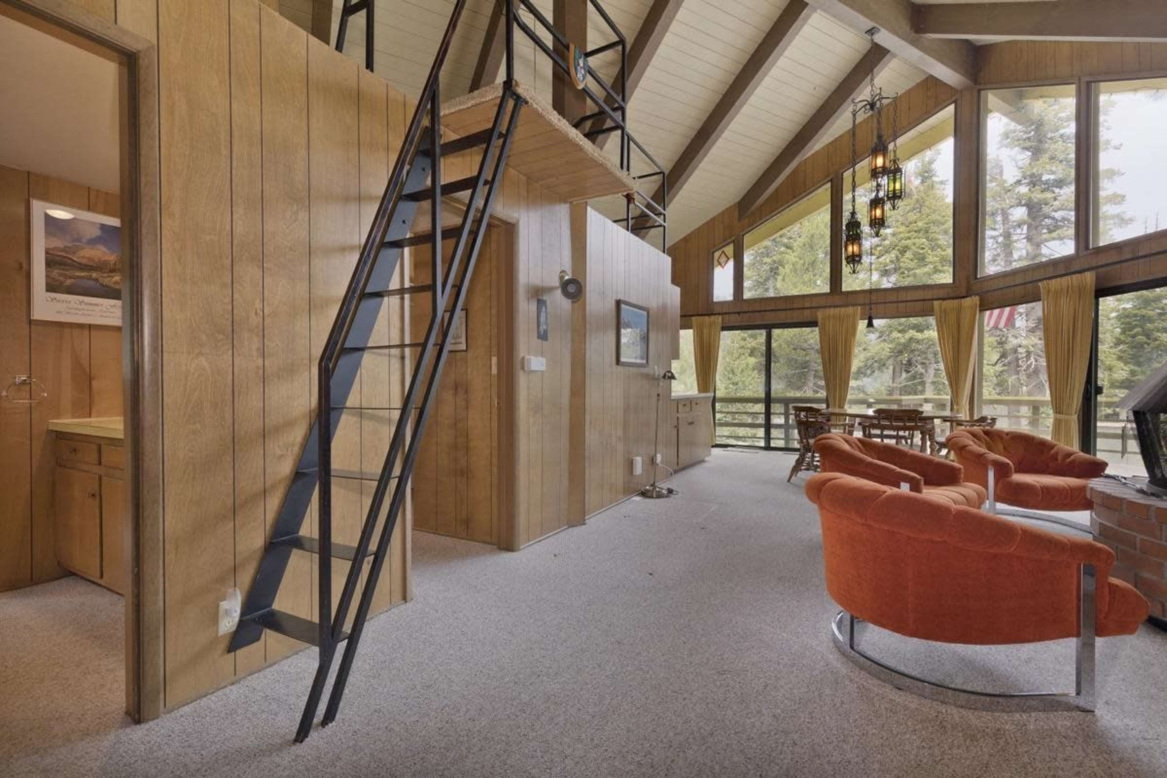 The interior of a cabin features wood-paneled walls, a raised loft accessible by a metal staircase, and seating arranged around a fireplace.