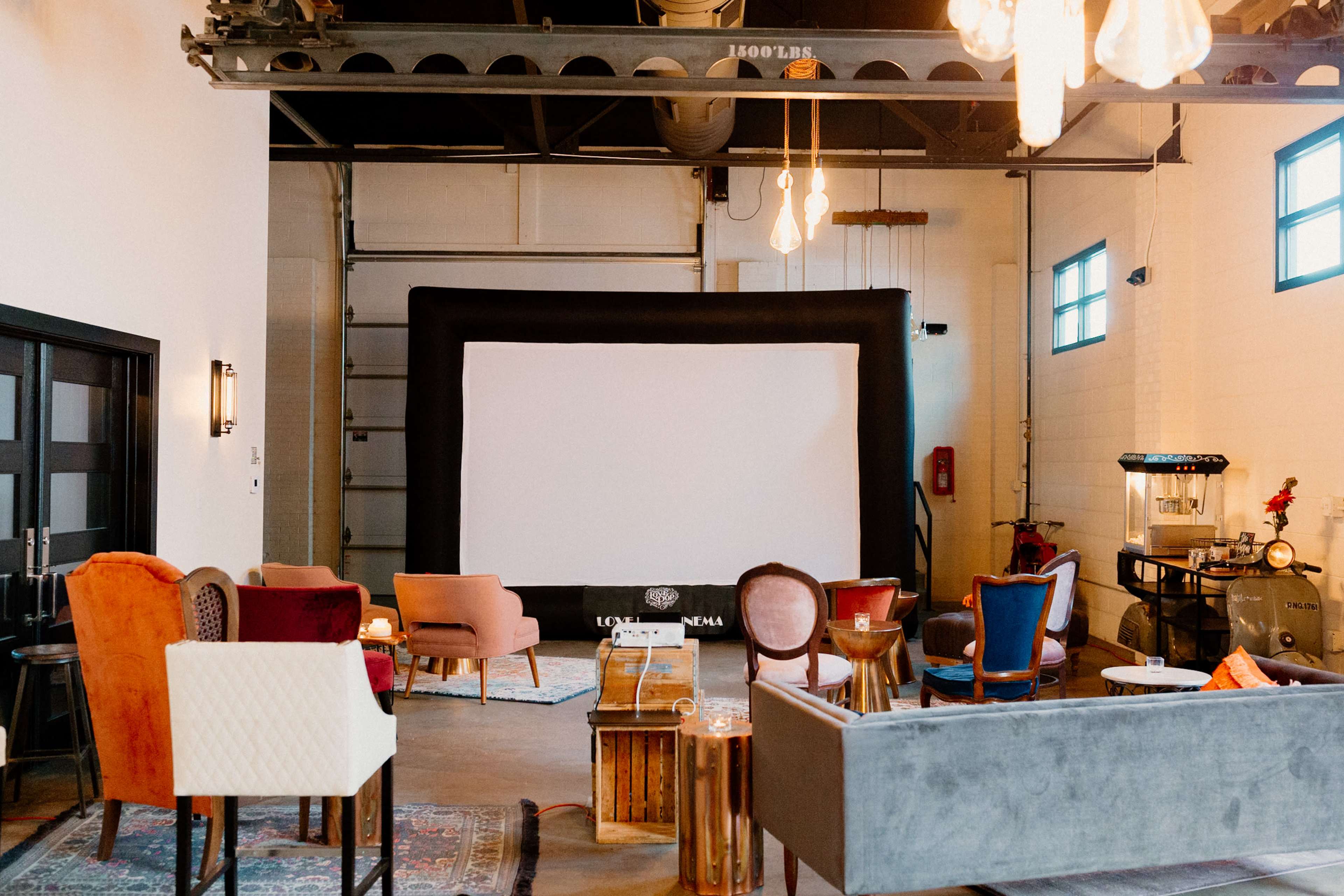 The image shows a spacious room with a large screen set up at one end, surrounded by various seating options including colorful chairs and couches.