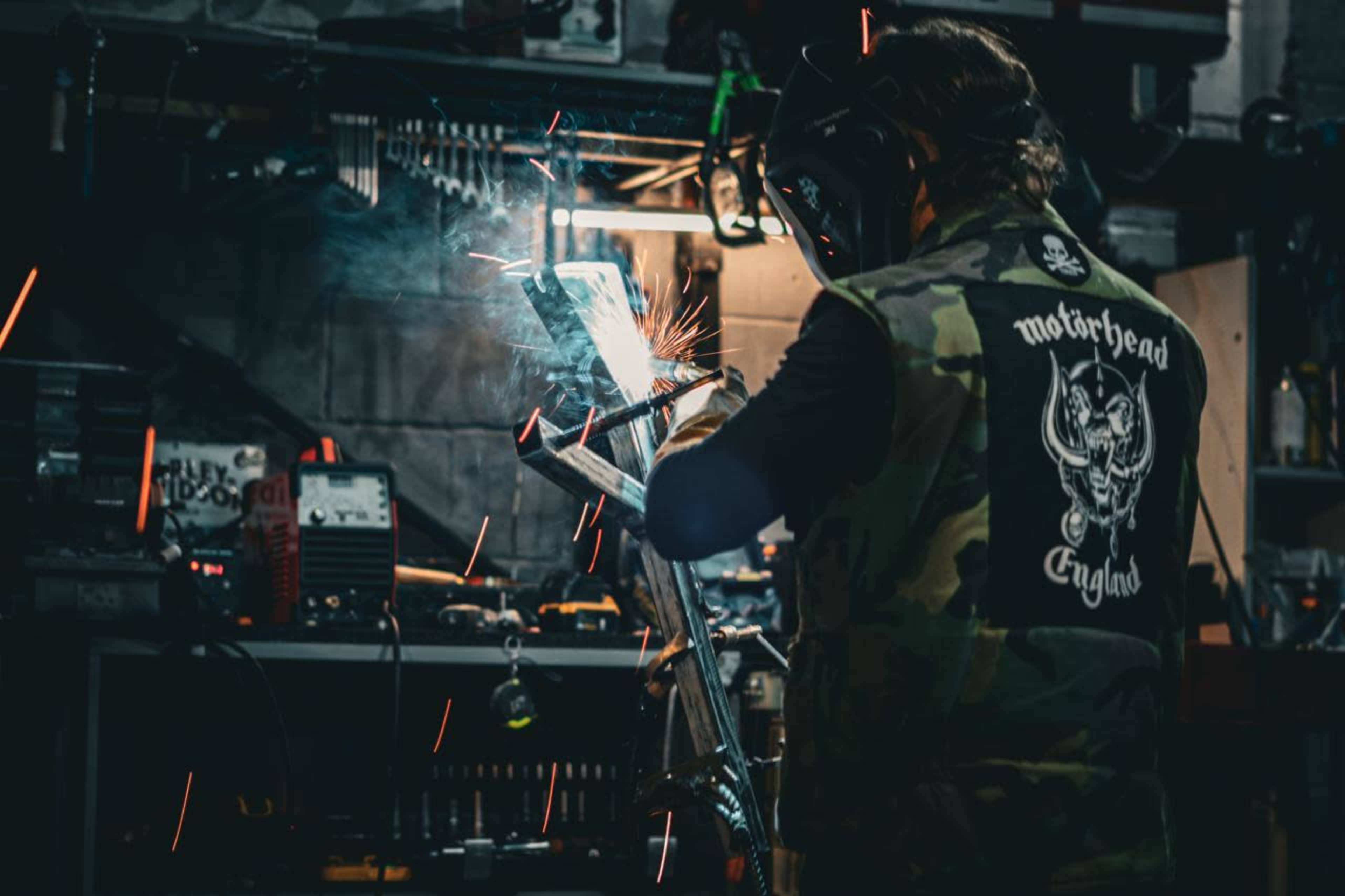 A person wearing a camouflage vest is welding a metal piece in a workshop, with sparks flying and various tools visible in the background.