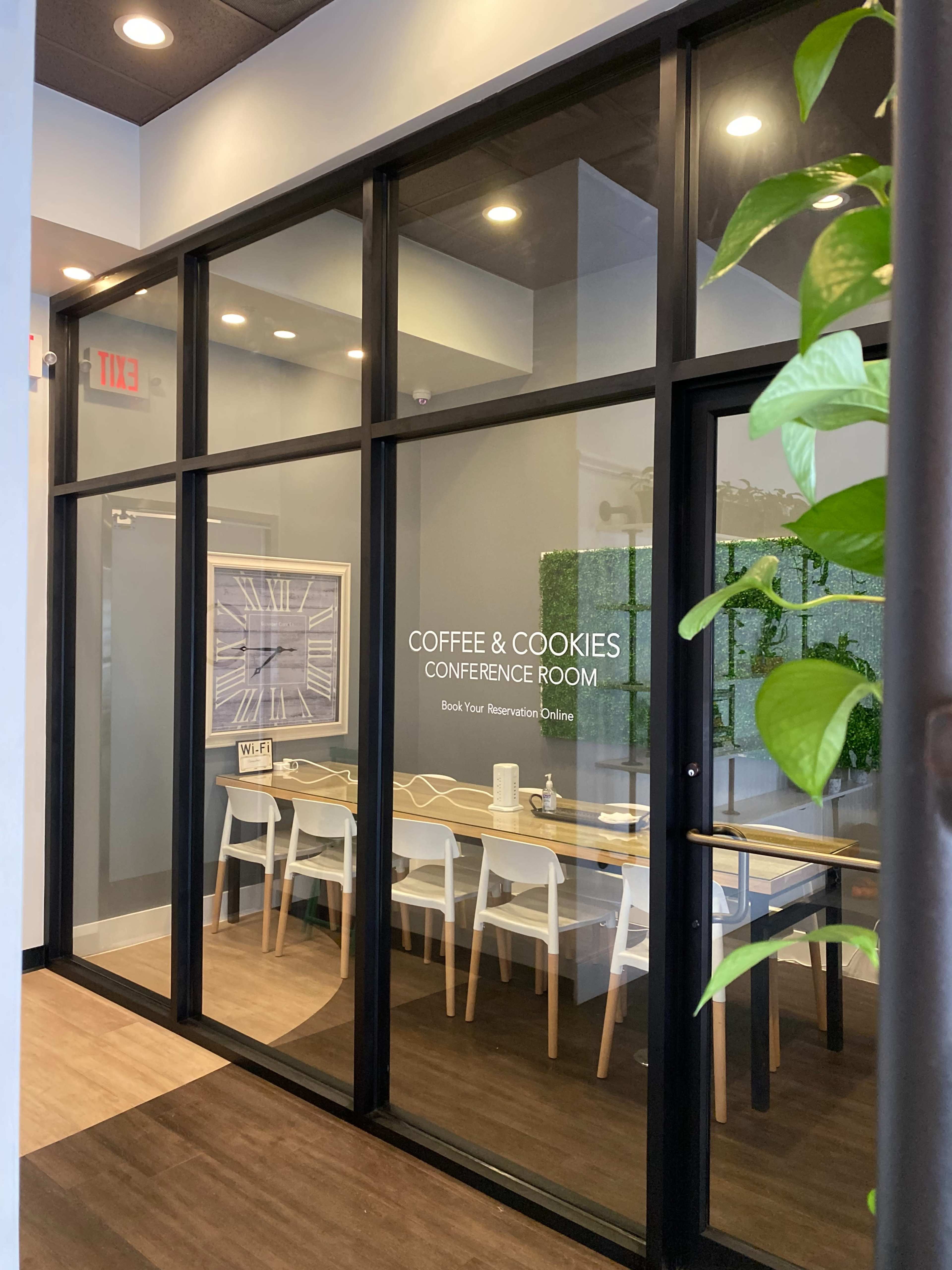 A glass-walled conference room titled "Coffee & Cookies" features a long table surrounded by white chairs and greenery decor.