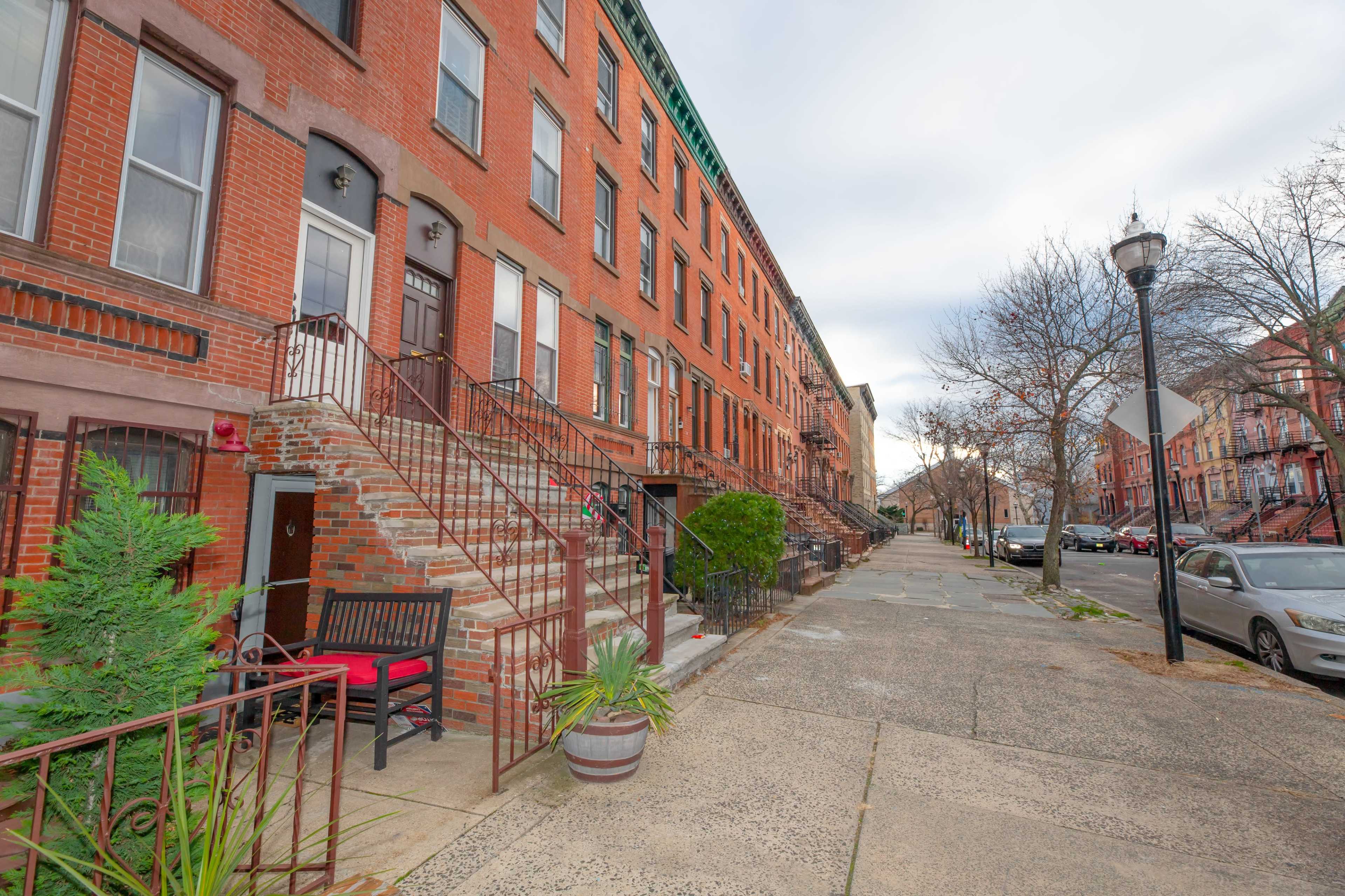 A residential street lined with brick townhouses, featuring stairs leading up to the front doors and a sidewalk with parked cars.