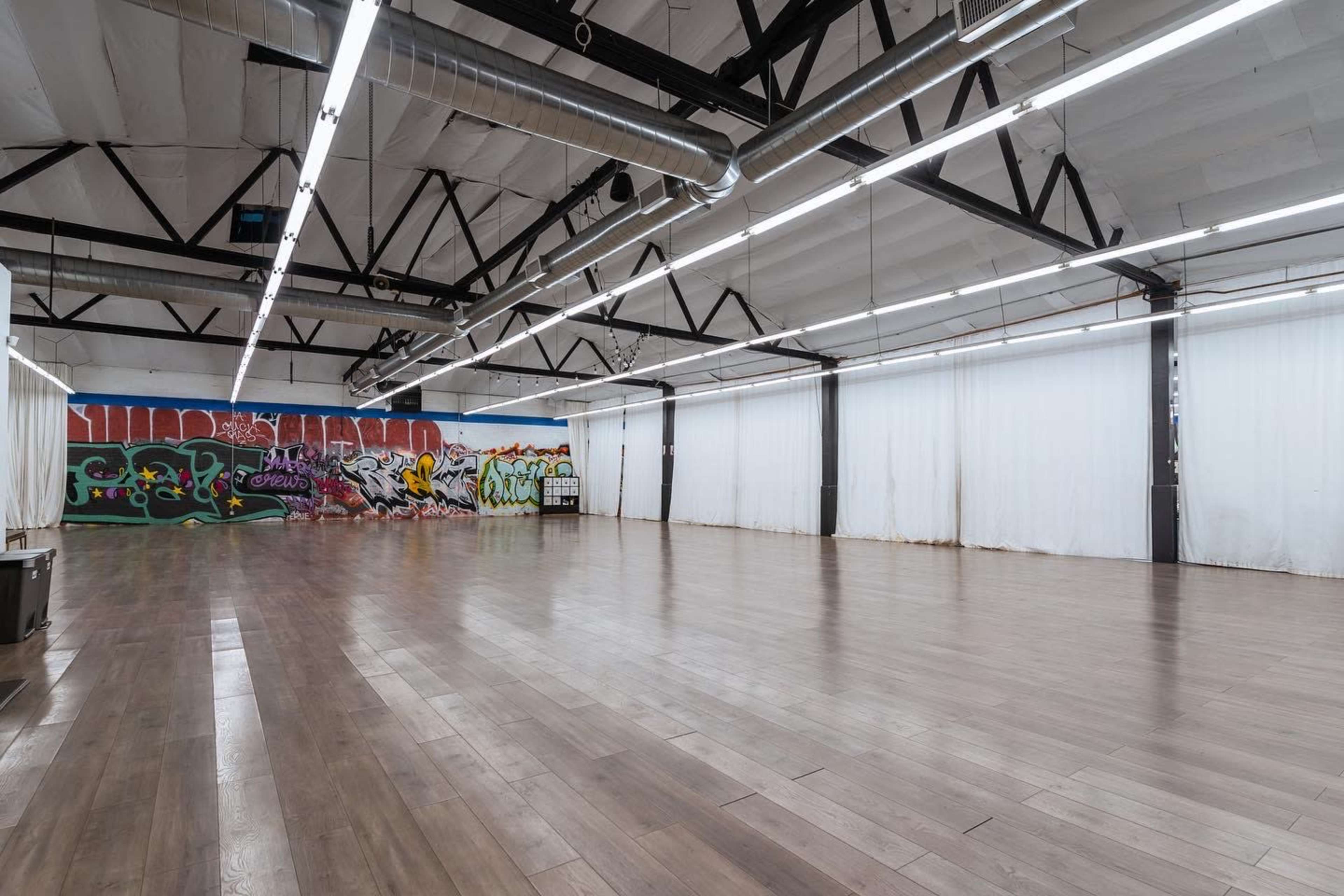 The image shows a spacious, empty industrial room with wooden flooring and a wall featuring colorful graffiti art.