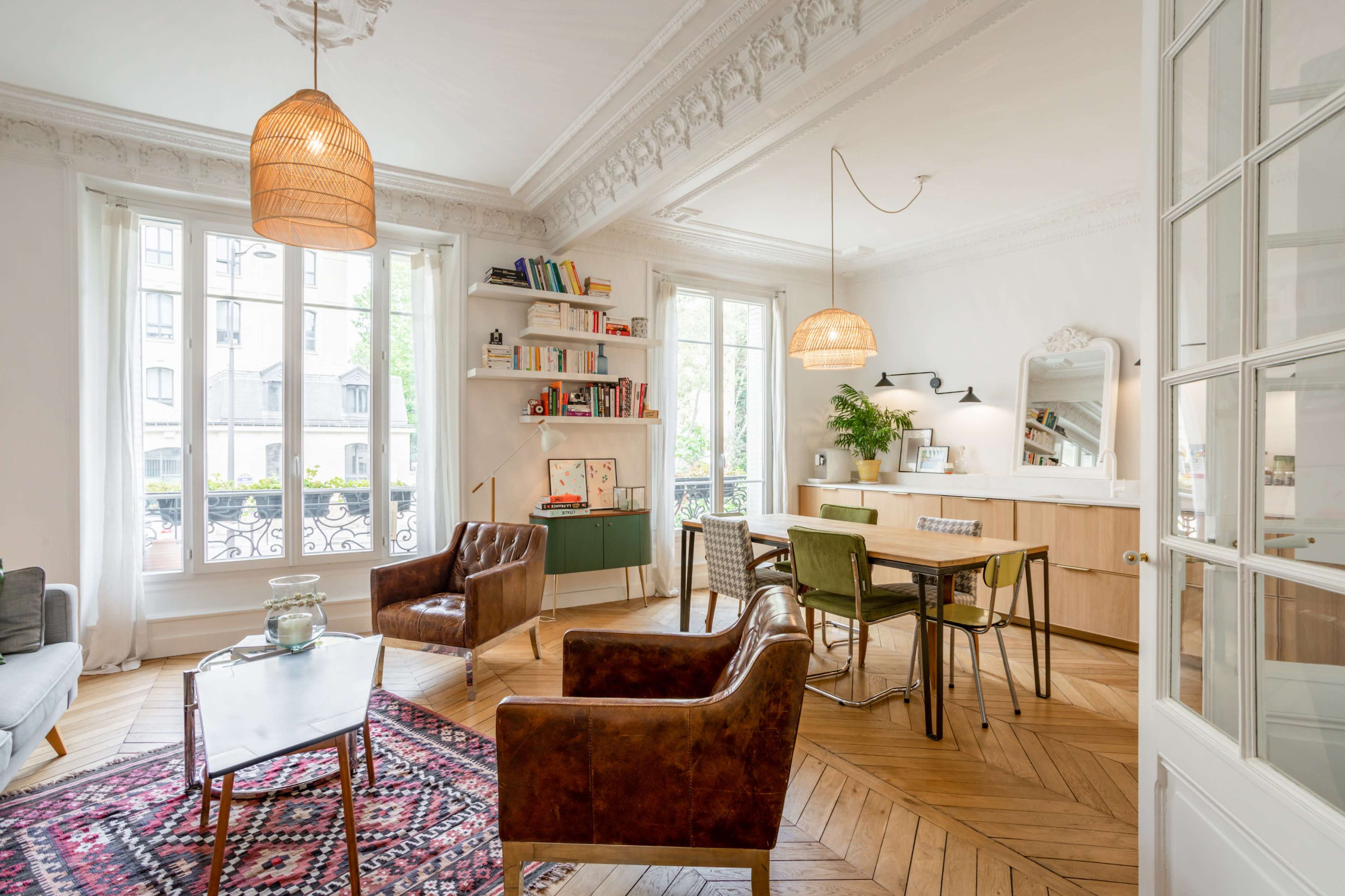 A well-lit living room with a mix of seating, a dining area, bookshelves, and large windows overlooking a street.